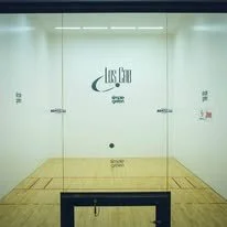 A squash court with glass walls, wooden floor, and a bench in front.
