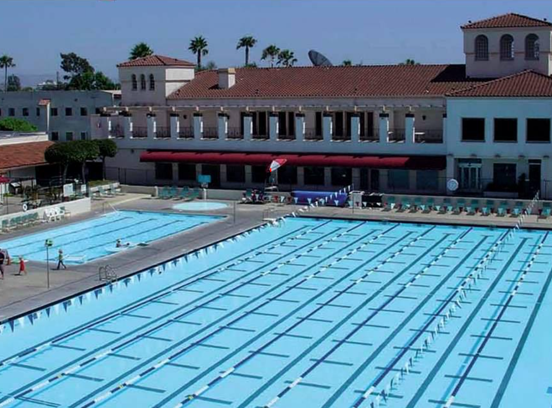 Outdoor swimming pool with clear blue water, empty lanes, and a few swimmers, surrounded by a deck with lounge chairs, umbrellas, and a building in the background with red-tiled roof and palm trees.