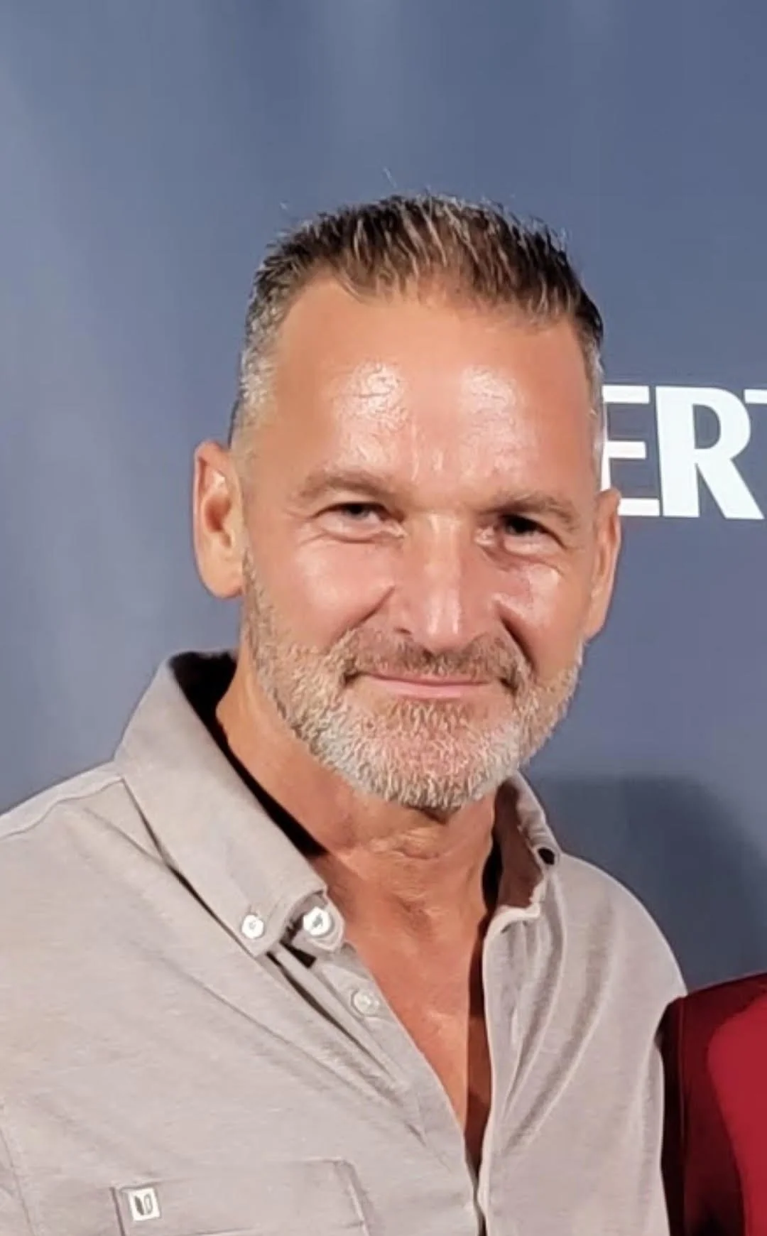 A middle-aged man with short hair and a beard, smiling, wearing a beige button-up shirt, standing in front of a gray backdrop.