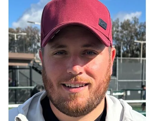 A young man with a beard wearing a red baseball cap, outdoors on a sunny day with blue sky and trees in the background.