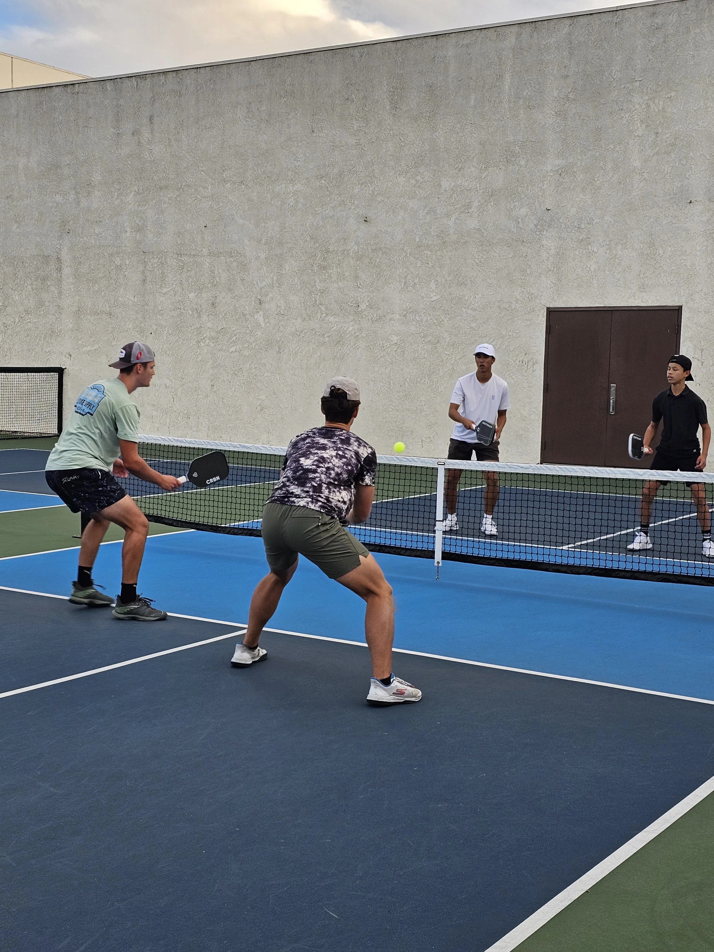 Four young men playing pickleball on a blue-colored court near a beige wall, with one active player in focus preparing to hit a yellow ball while three others watch.