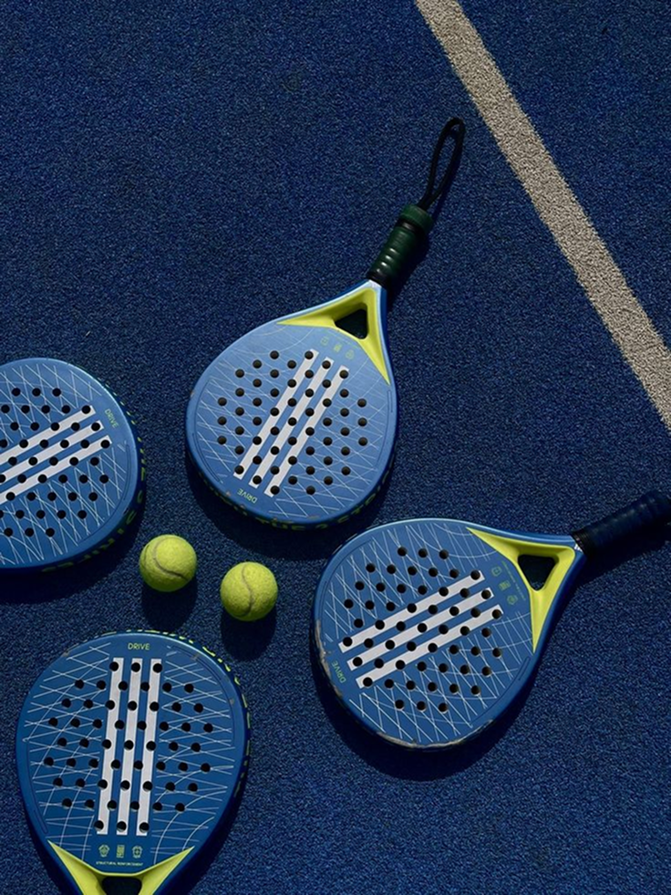 Two blue padel rackets with yellow accents and two yellow tennis balls on a blue tennis court.