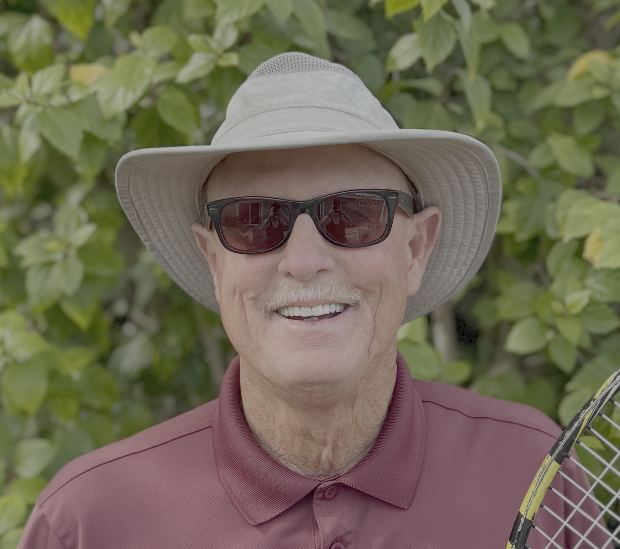 Close-up of an elderly man wearing a wide-brimmed hat, sunglasses, and a maroon polo shirt, holding a tennis racket, standing in front of green leafy bushes.