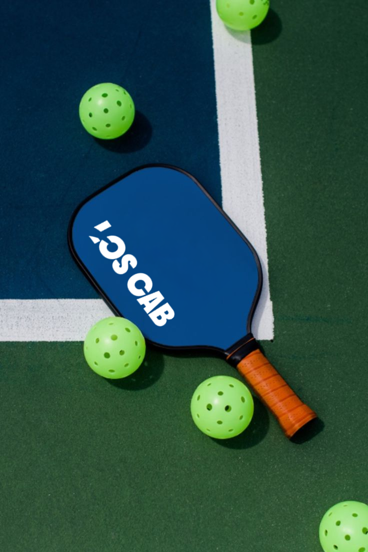 A blue pickleball paddle with the words 'SMSC' on it, surrounded by green perforated pickleballs on a blue and green tennis court.