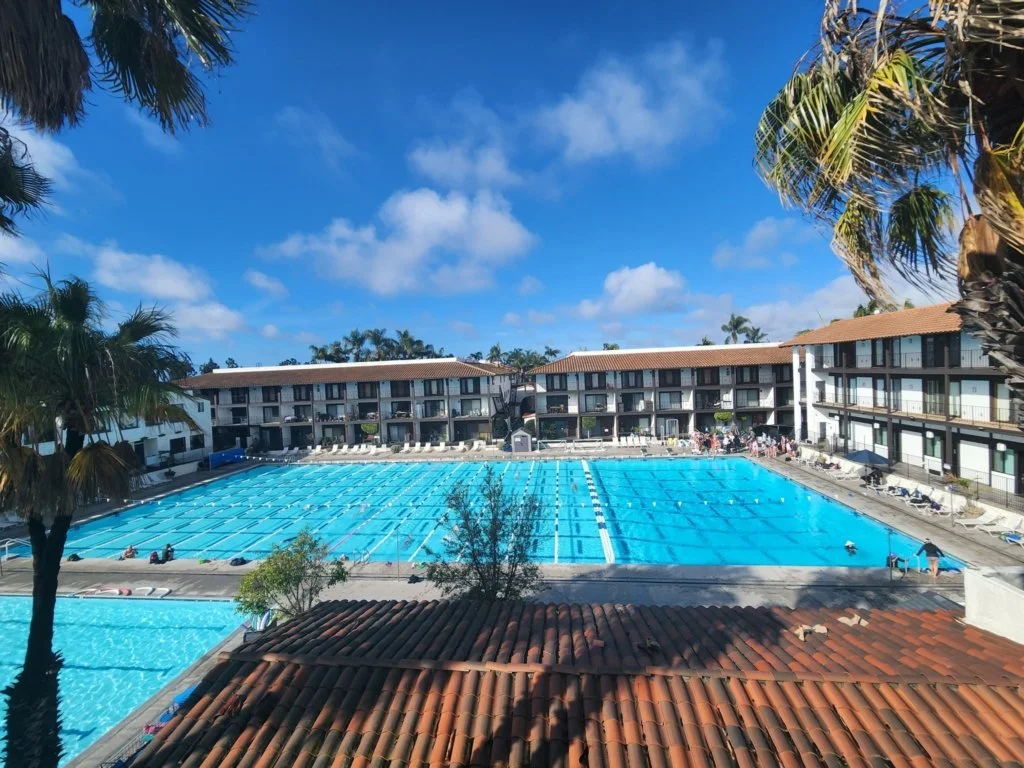 View of a large outdoor swimming pool at a hotel with multiple levels of balconies, surrounded by palm trees and lounge chairs, under a blue sky with some clouds.