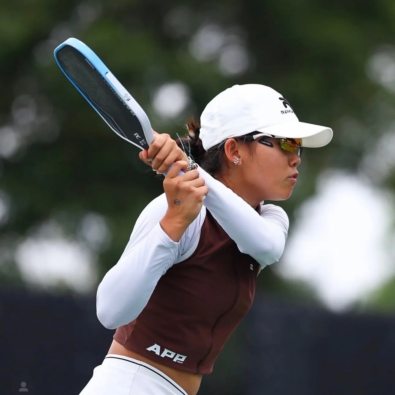 Female golfer wearing sunglasses, a white cap, and a long-sleeve shirt, preparing to swing her golf club outdoors.