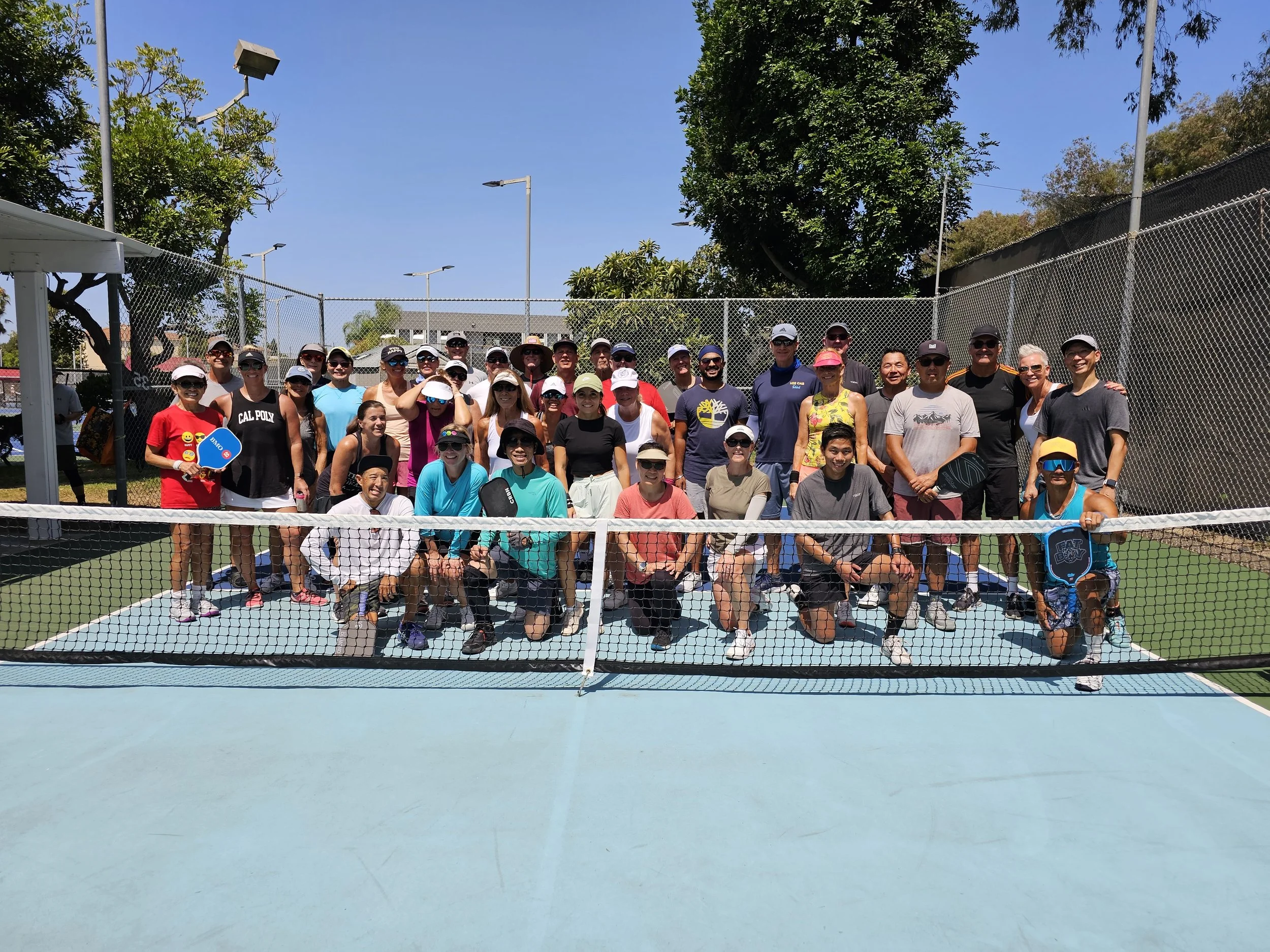 Group of people posing for a photo on a tennis court under sunny weather, with trees and a chain-link fence in the background.