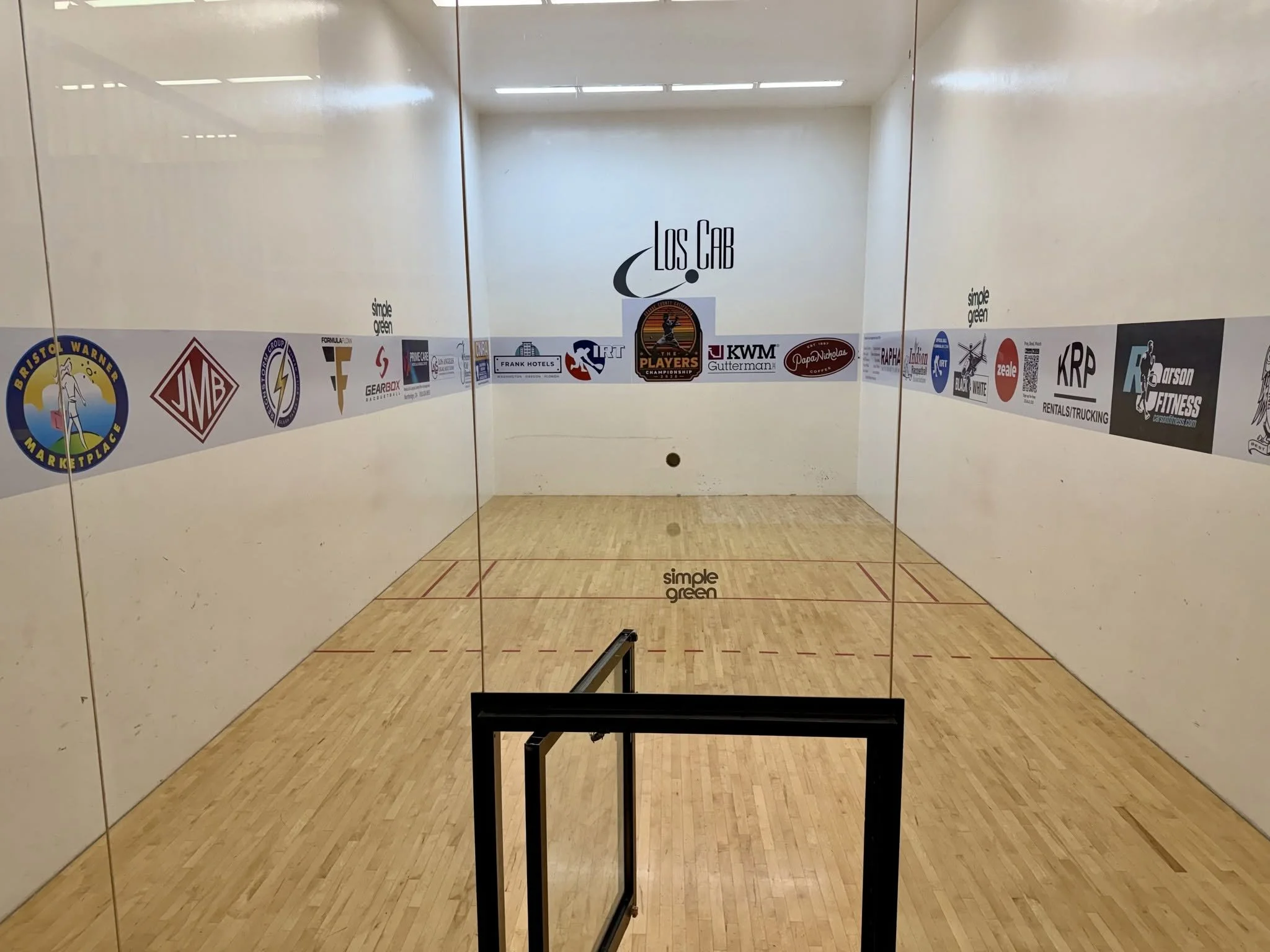 A squash court with white walls, wooden flooring, and a glass back wall with alphabetical and sponsor logos, including 'Los Cab' in the center, taken from the perspective of a person standing outside the court.