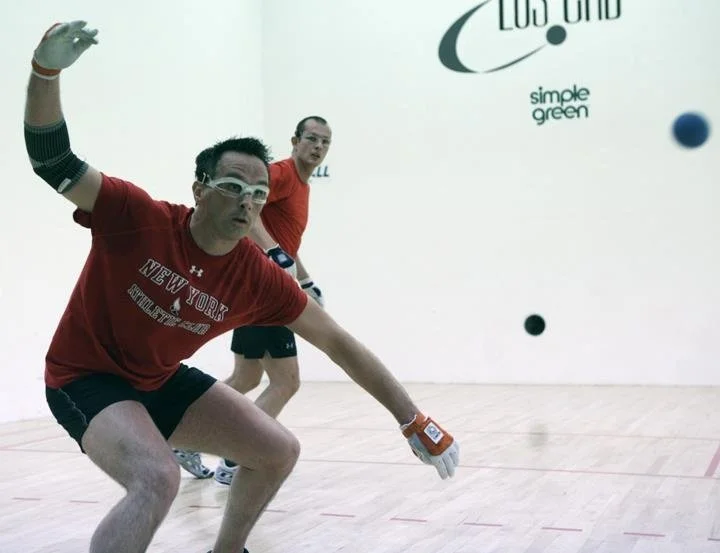 Two men playing racquetball in an indoor court, one reaching to hit a racquetball with a racquet, while the other stands behind observing.