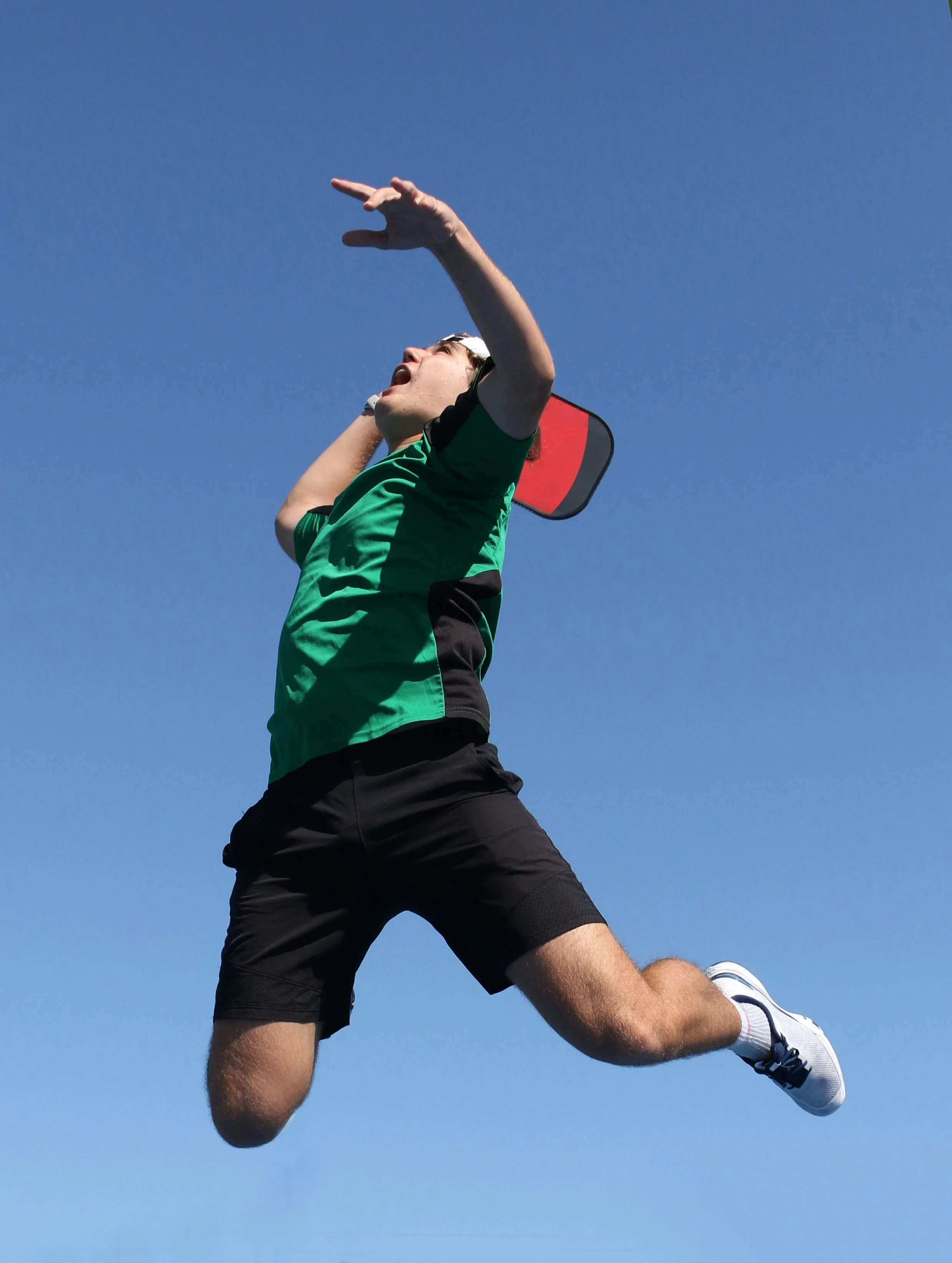 Young man mid-air against a clear blue sky, jumping or falling with his mouth open and one arm raised.