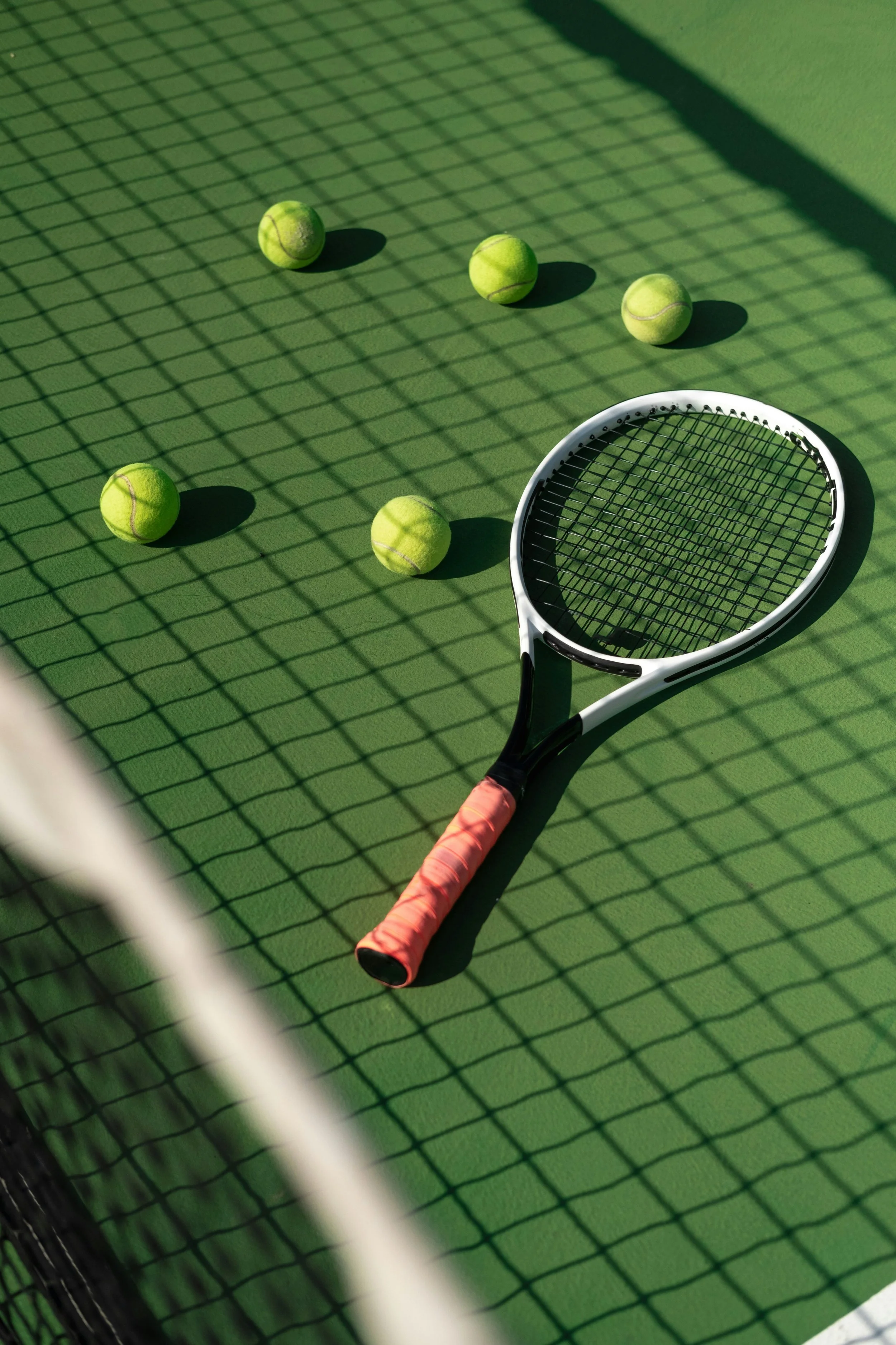 Six tennis balls, a tennis racket, and a tennis net shadow on a green tennis court