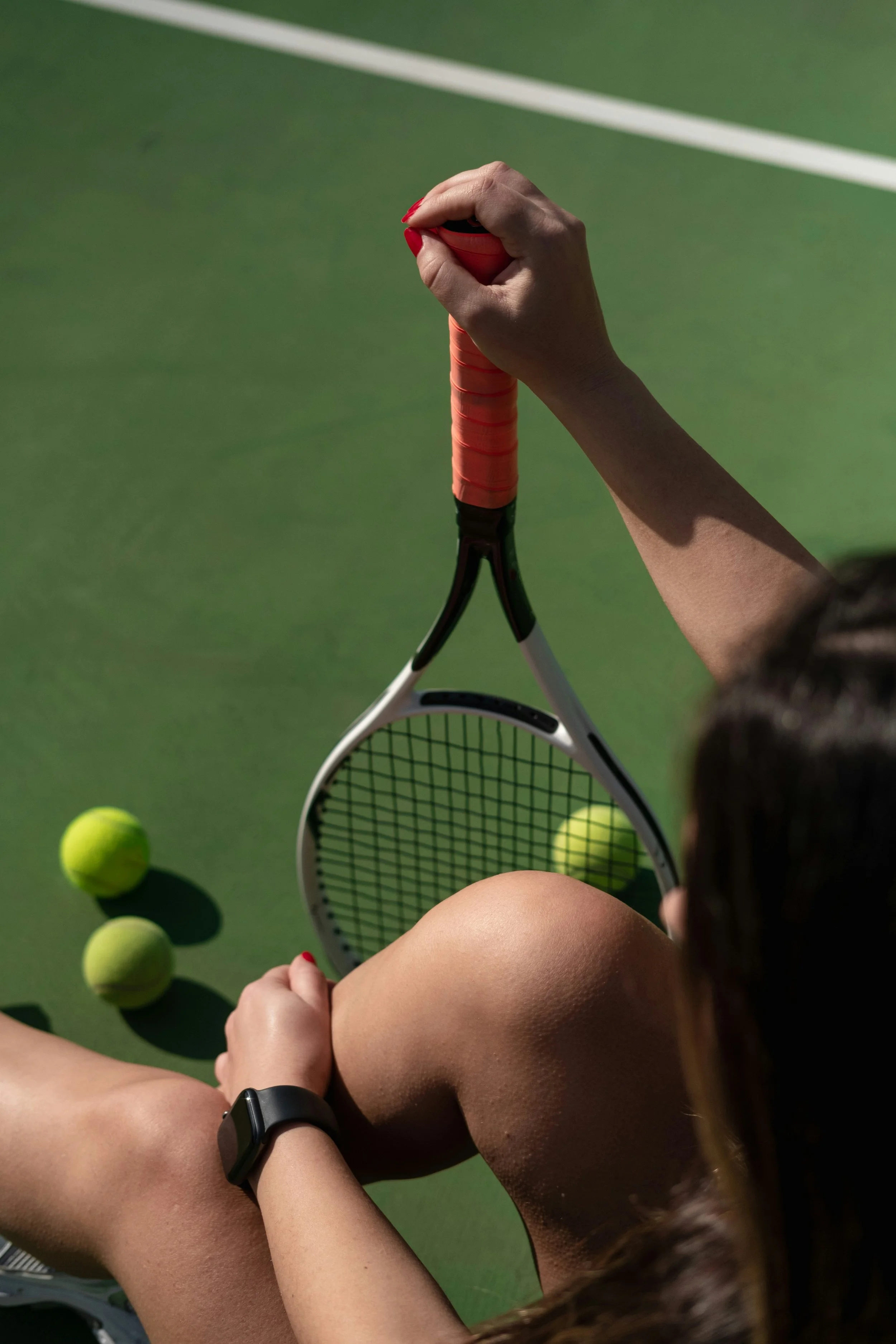 A person sitting on a tennis court holding a tennis racket with tennis balls on the ground around them.