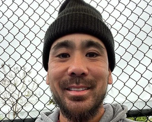 Close-up of a man with a beard and mustache wearing a black beanie and gray hoodie, smiling in front of a chain-link fence with trees and cloudy sky in the background.