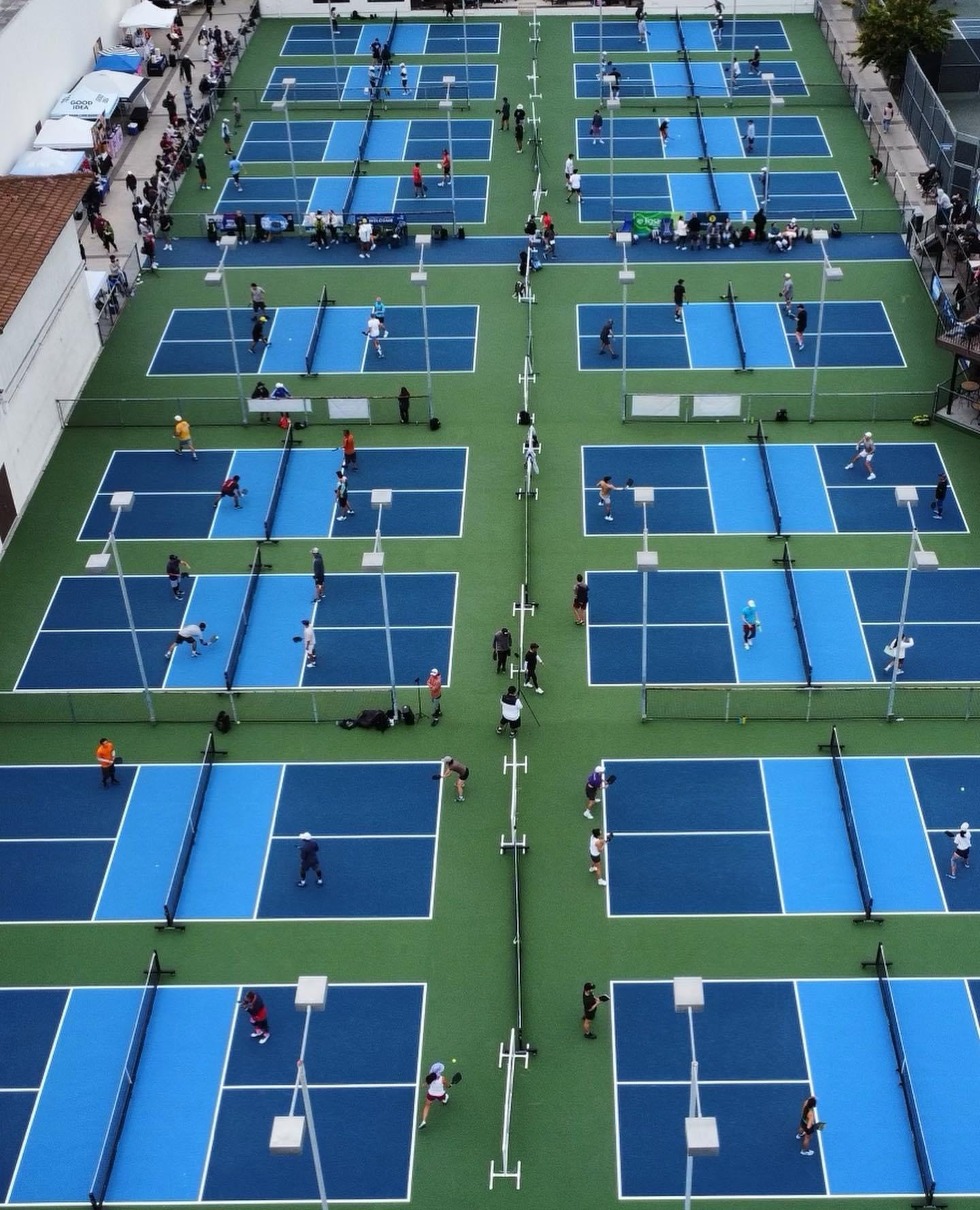 Bird's eye view of multiple outdoor blue tennis courts with players and spectators, separated by fences, on a green surface.