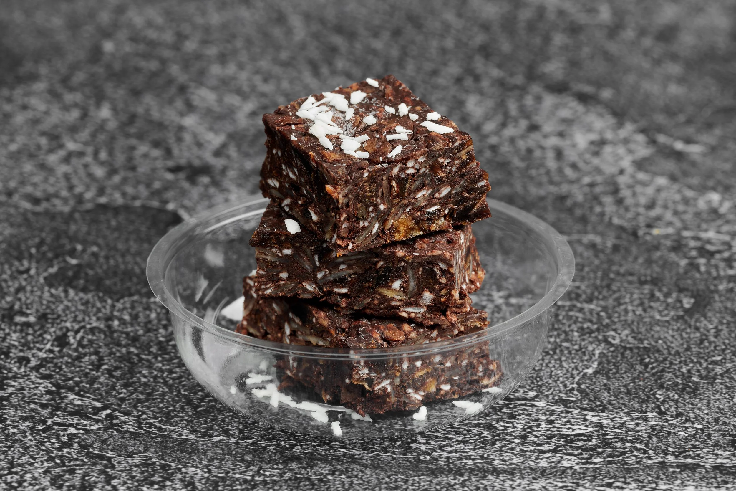 Stack of chocolate bark with nuts and white flakes in a clear glass bowl on a speckled black surface.