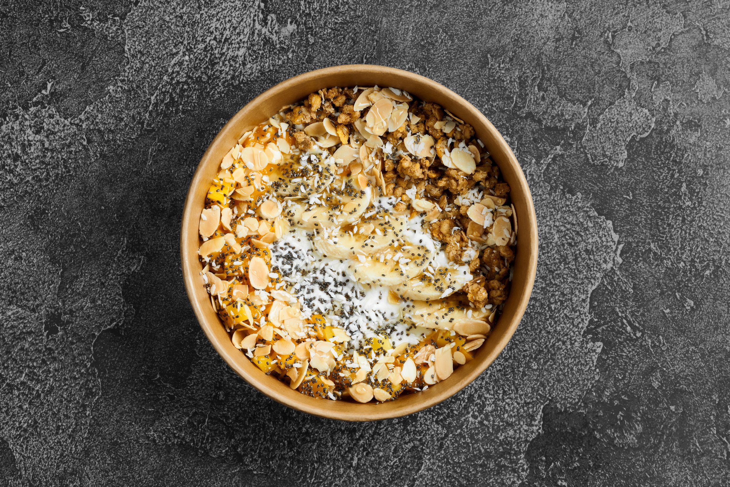 A bowl of food containing sliced bananas, vanilla pudding with black chia seeds, almond slices, and crumbled cookies on a dark, textured surface.
