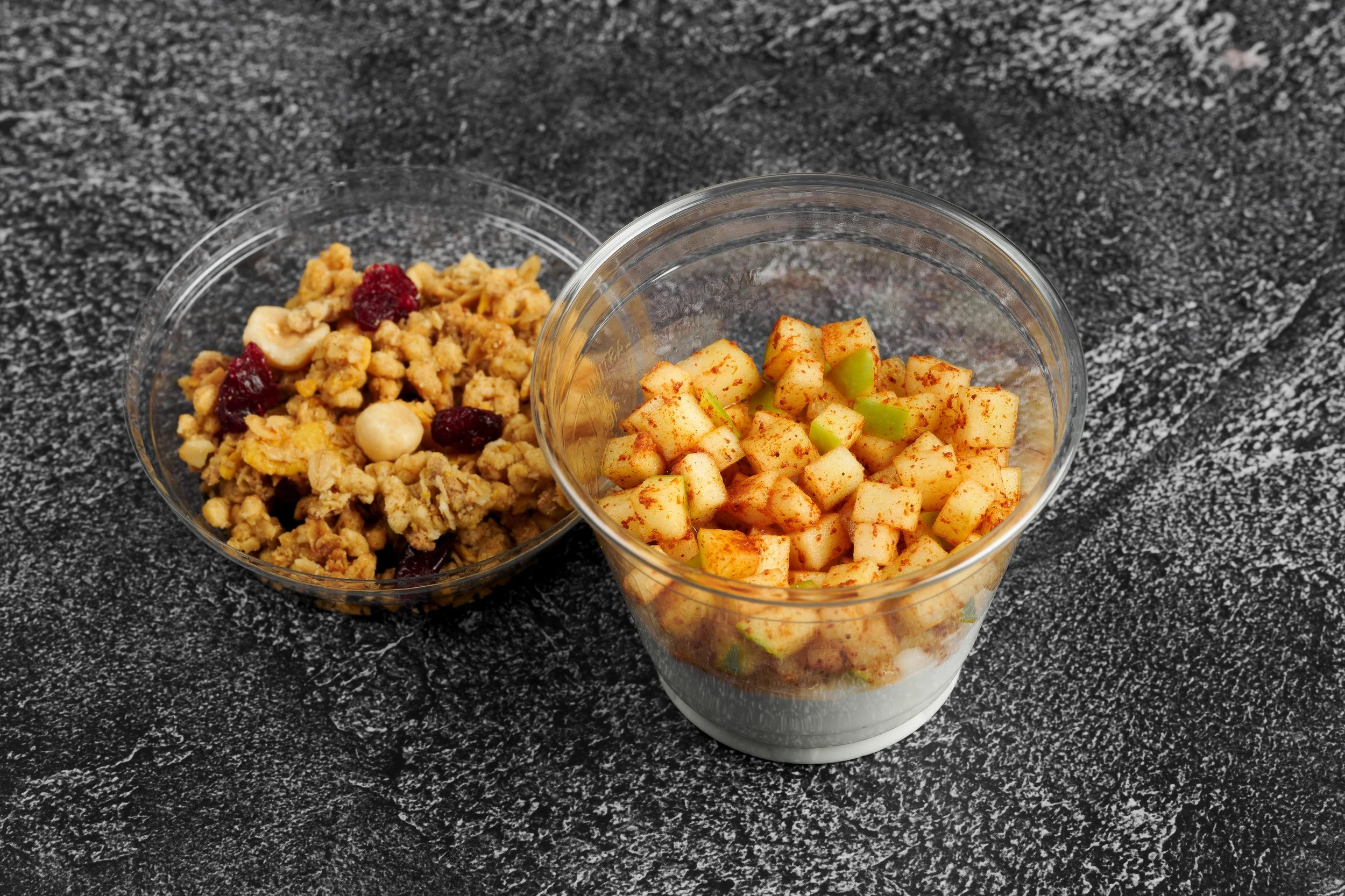 Two clear plastic cups on a dark gray speckled countertop, one filled with cooked fruit and the other with chopped fruit seasoned with red spice, possibly paprika or chili powder.