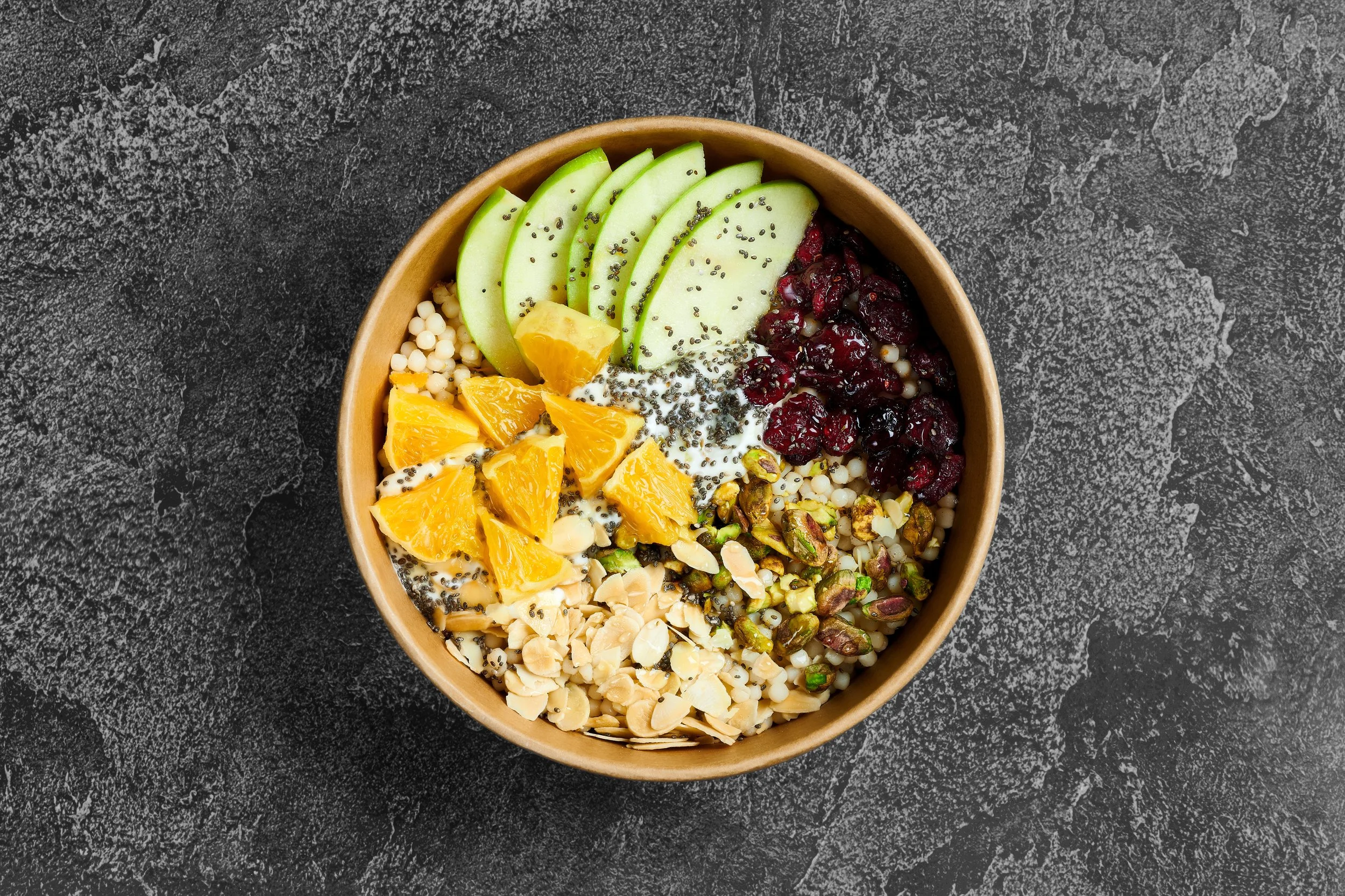 Bowl of smoothie topped with sliced green apples, orange segments, dried cranberries, seeds, and nuts on a black textured surface.