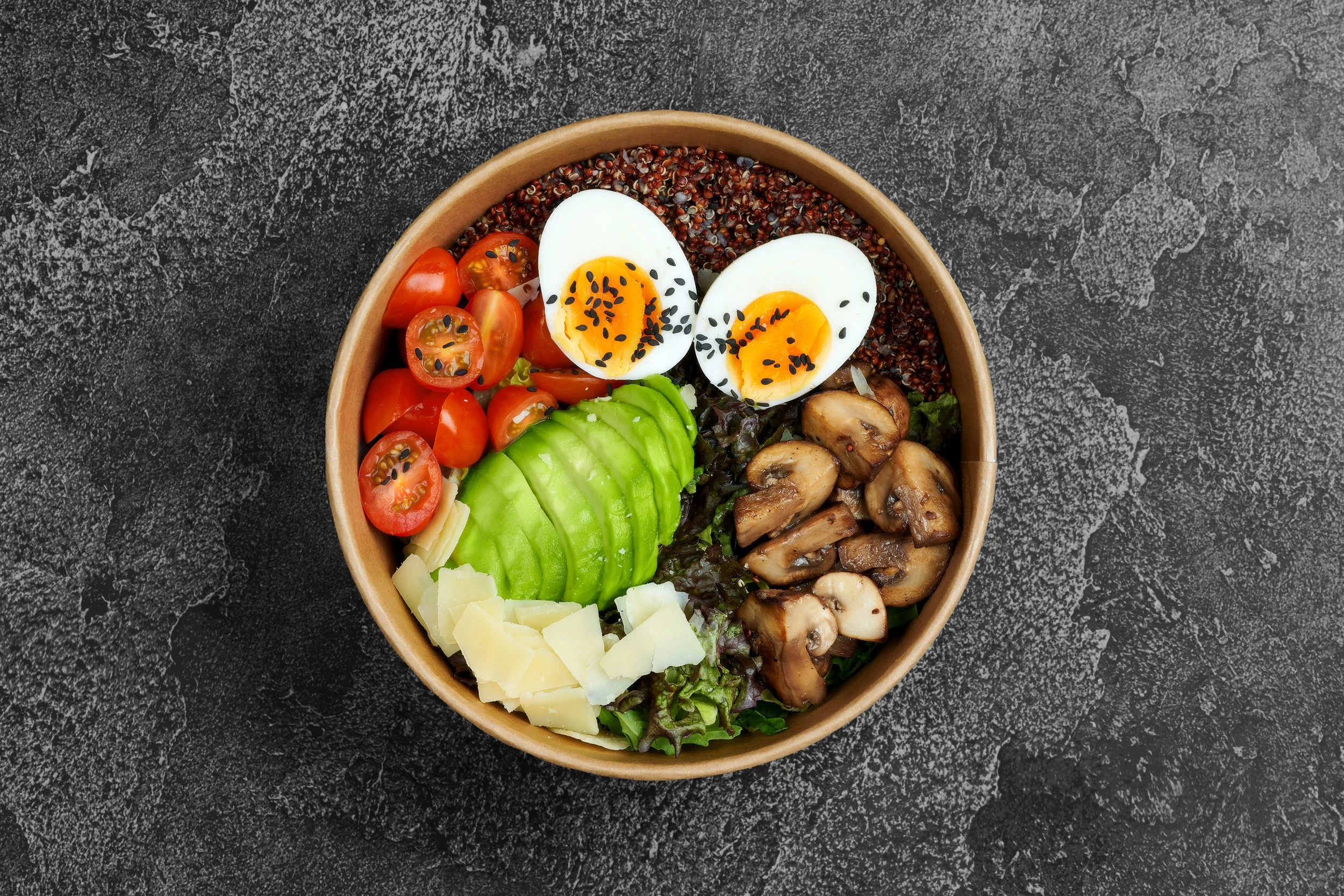 A bowl containing a salad with sliced avocado, cherry tomatoes, boiled eggs sprinkled with black sesame seeds, sautéed mushrooms, cheese shavings, and cooked quinoa, on a dark textured surface.