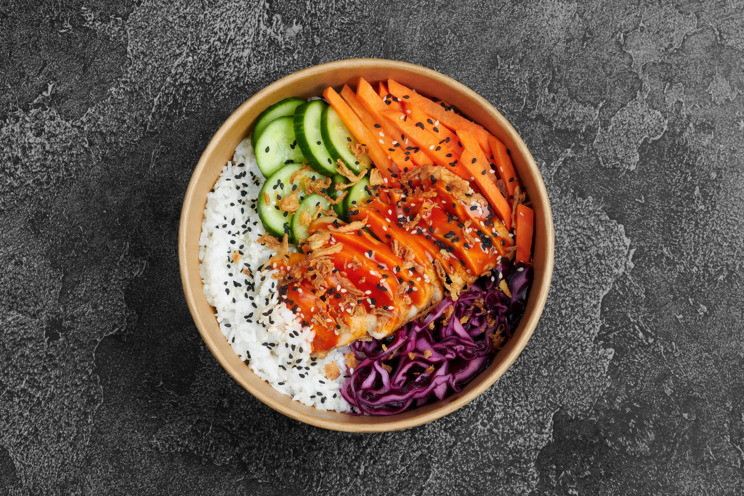 Bowl of rice with sliced cucumber, shredded carrots, purple cabbage, and sauce, garnished with black sesame seeds and onion flakes on a dark textured surface.