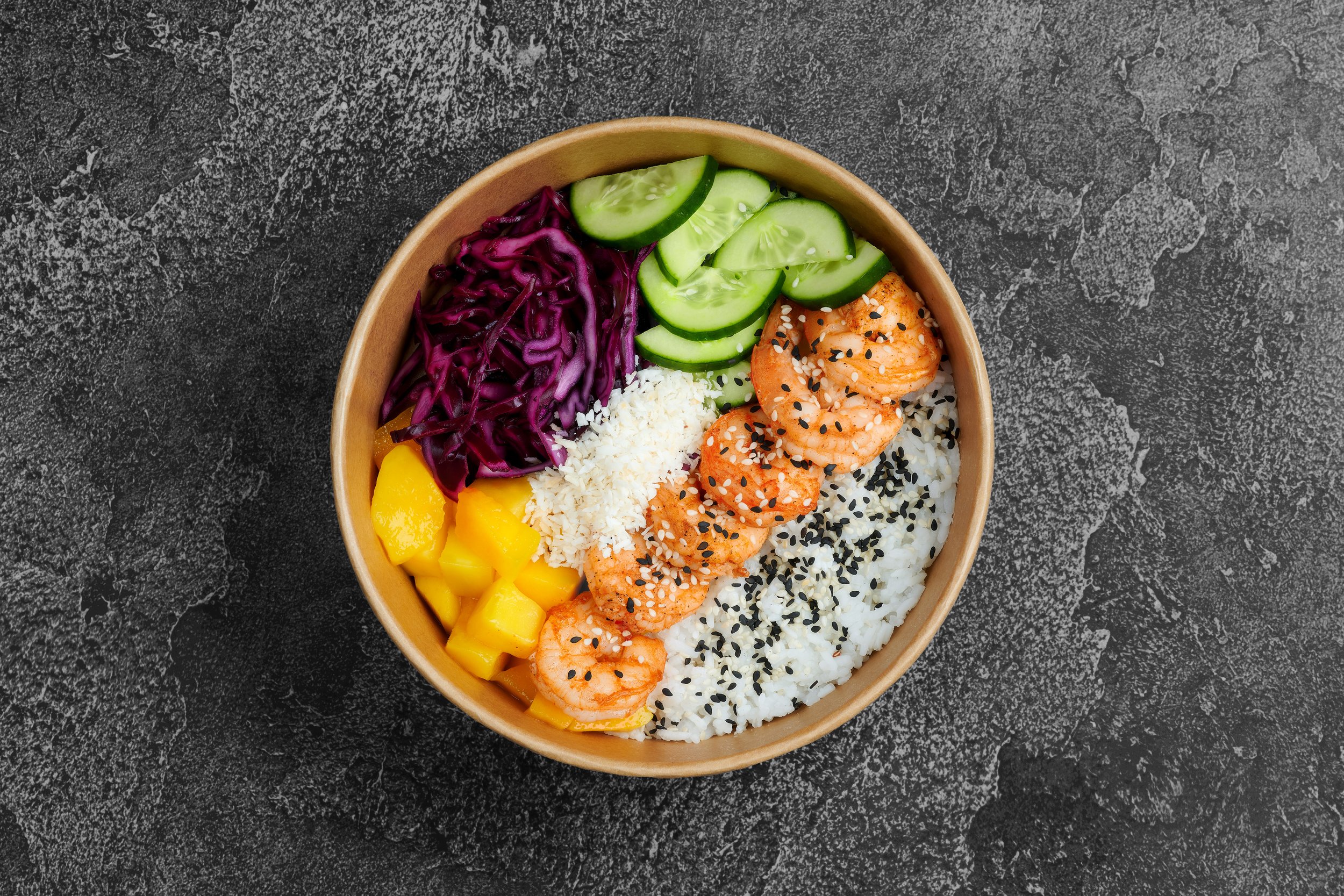 Bowl of Asian-inspired rice bowl with shrimp, cucumber slices, shredded purple cabbage, diced mango, white shredded cheese, and black sesame seeds, on a dark textured surface.