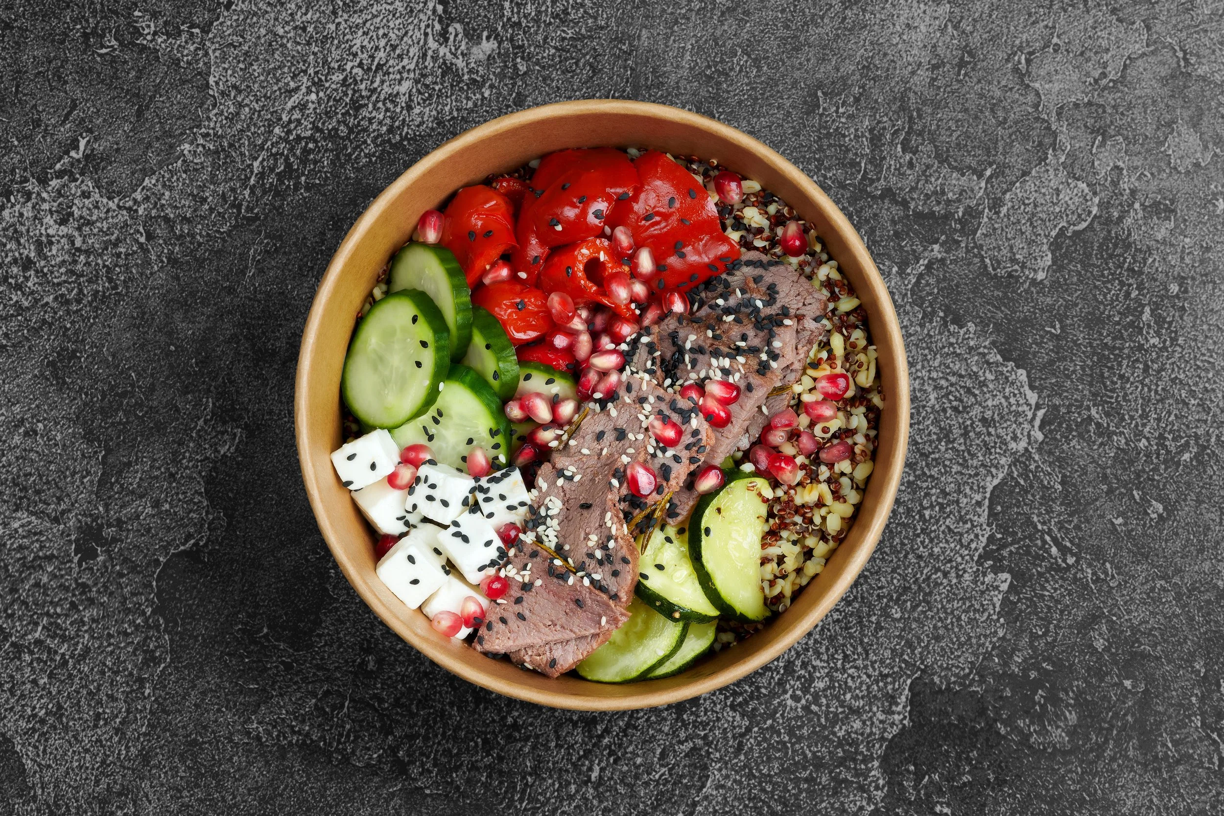 A wooden bowl filled with a variety of salad ingredients including sliced cucumbers, cherry tomatoes, cooked quinoa, diced feta cheese, and slices of beef topped with sesame seeds and pomegranate seeds, all placed on a dark textured surface.