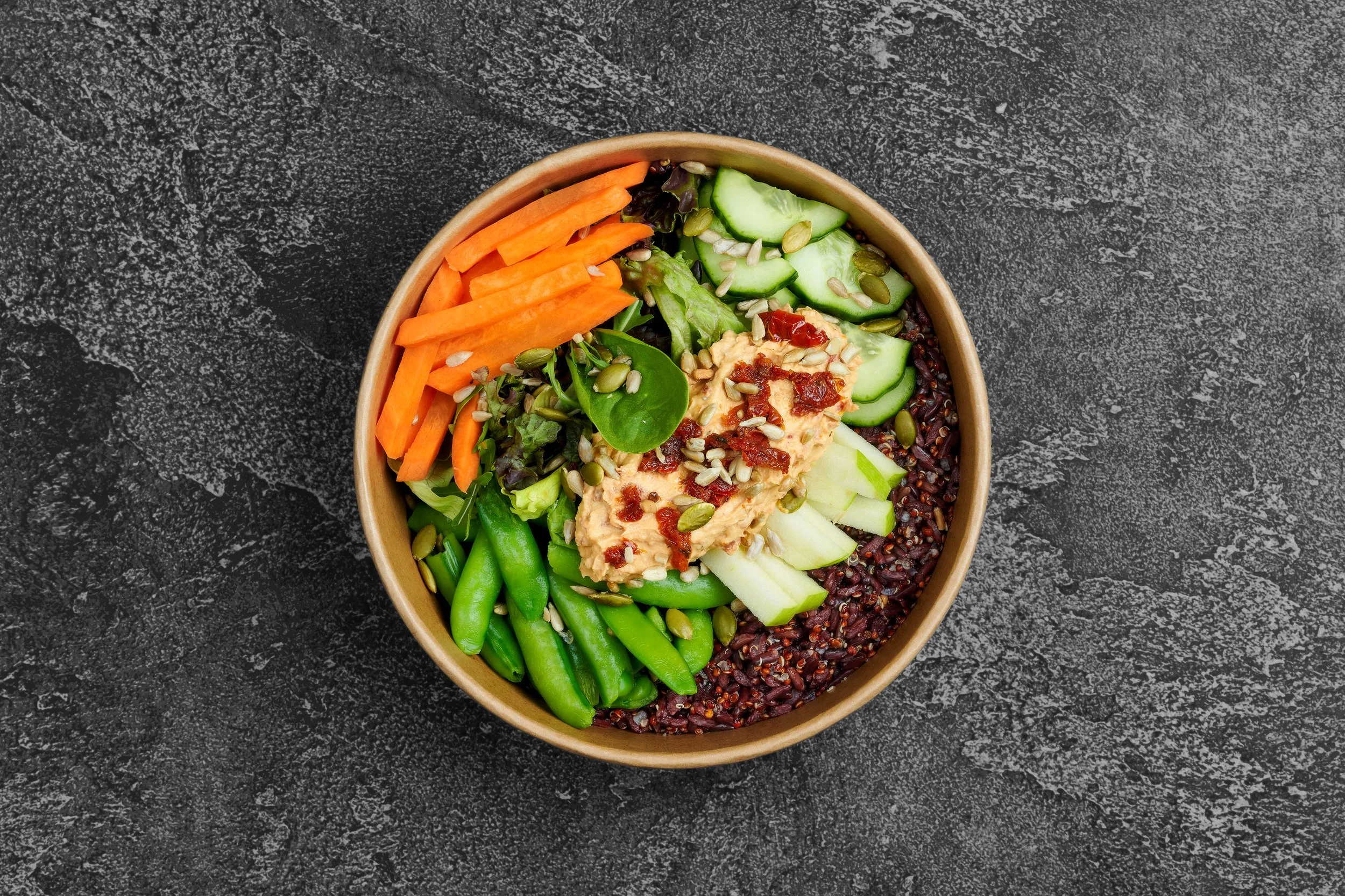 A bowl filled with a variety of fresh vegetables, including sliced carrots, cucumbers, leafy greens, green beans, and a dollop of hummus topped with sun-dried tomatoes and sunflower seeds, sitting on a dark textured surface.