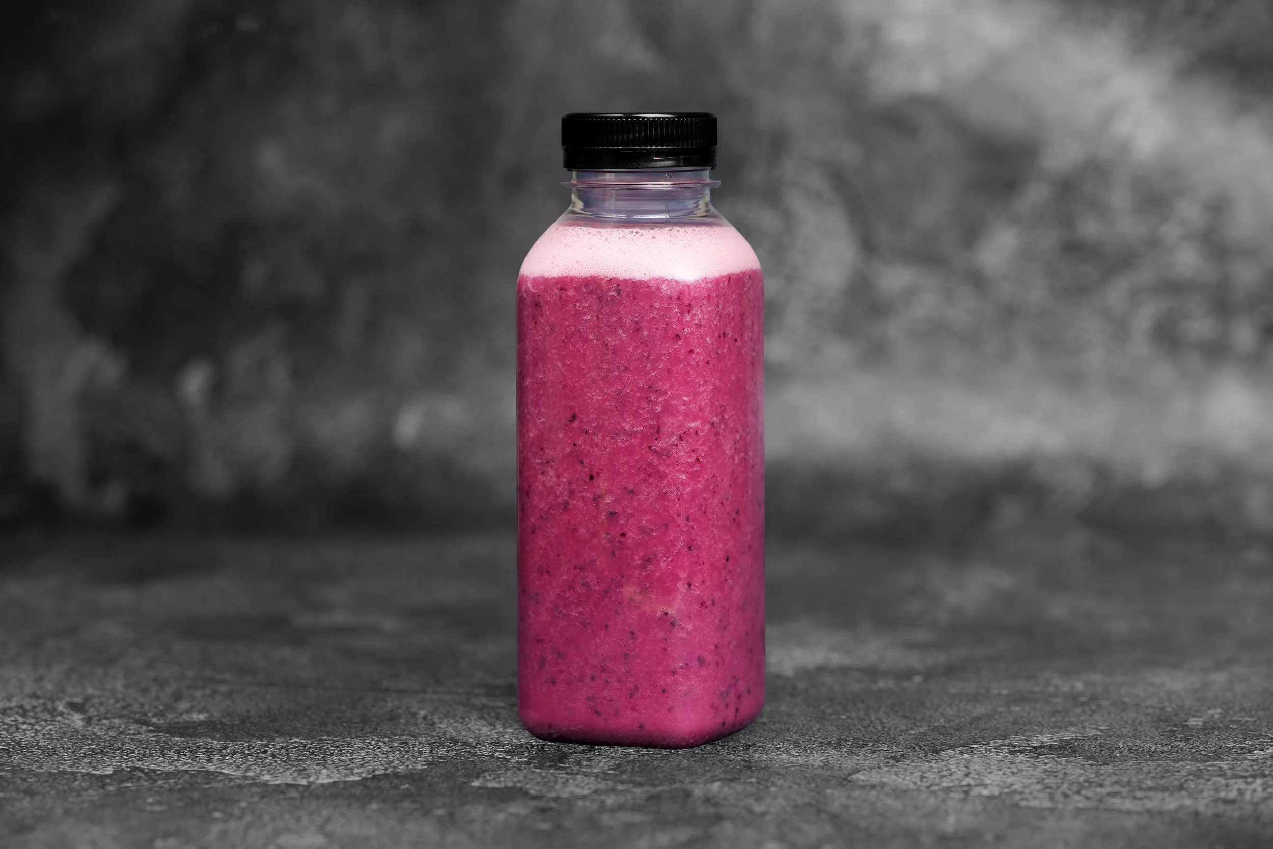 A pink smoothie in a clear plastic bottle with a black screw cap, placed on a gray textured surface with a dark, blurry background.
