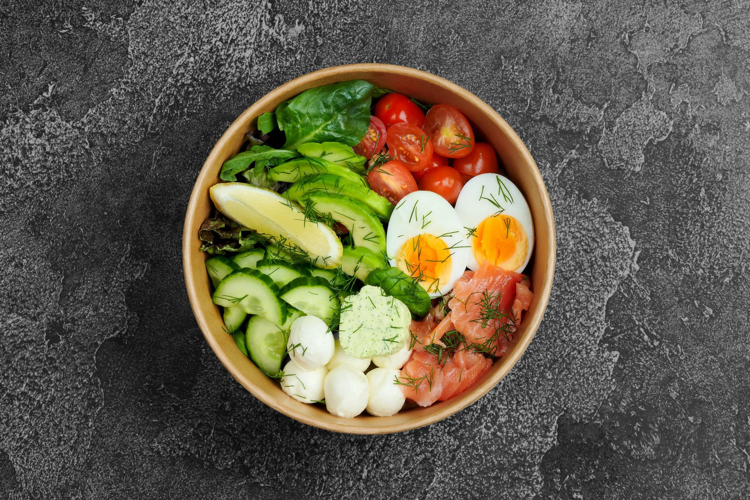 A wooden bowl filled with salad ingredients including cherry tomatoes, sliced cucumbers, boiled eggs, avocado slices, lettuce, and dill, placed on a dark textured surface.