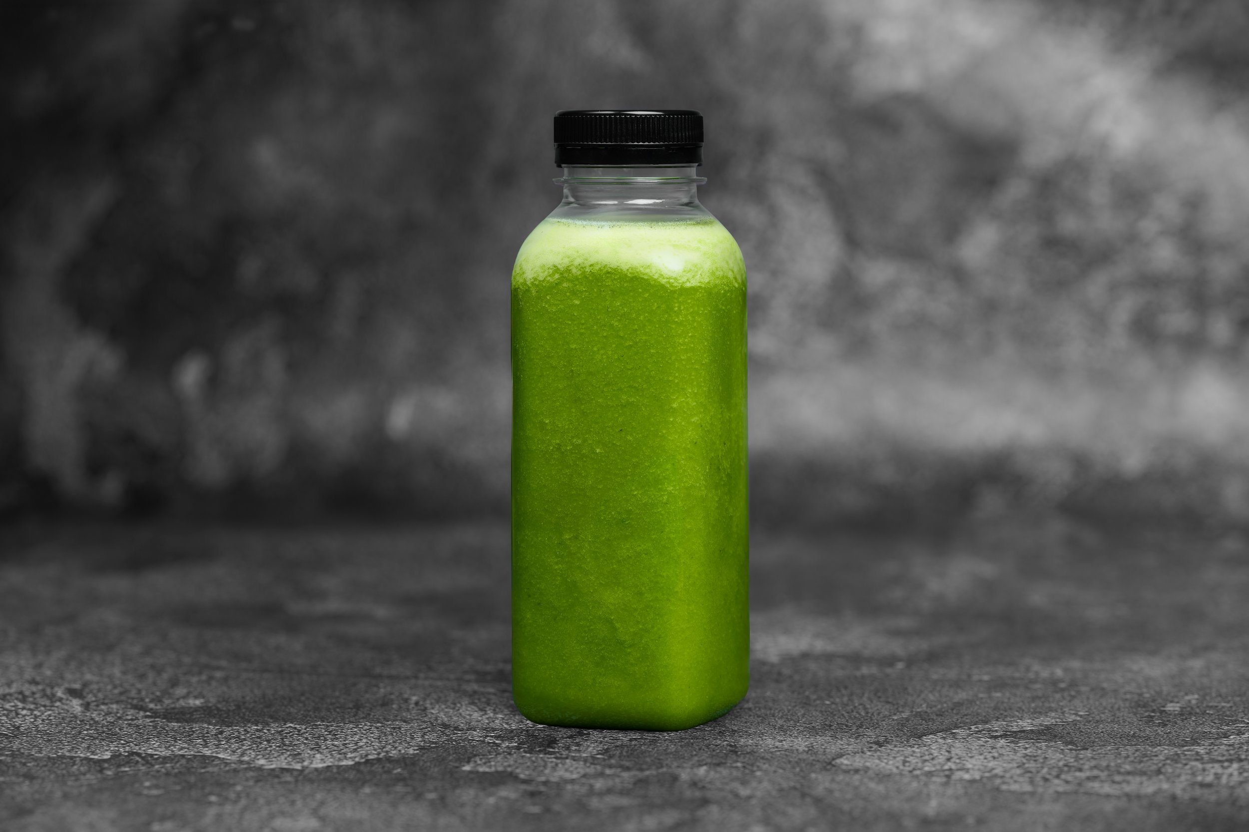 A clear glass bottle with a black cap, filled with a vibrant green liquid, on a textured dark gray surface with a blurred gray background.