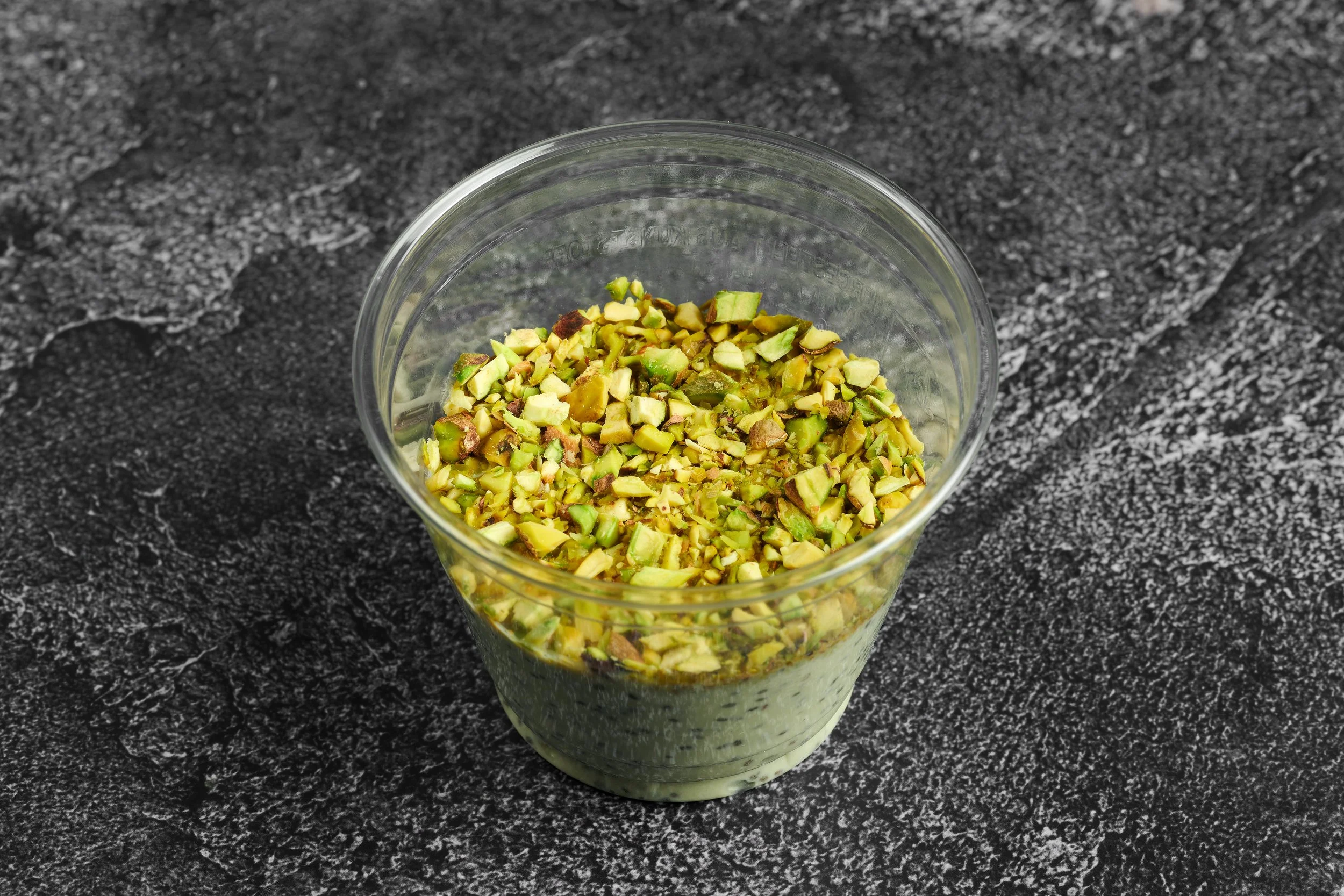 A clear glass cup filled with chopped pistachios on a black textured surface.