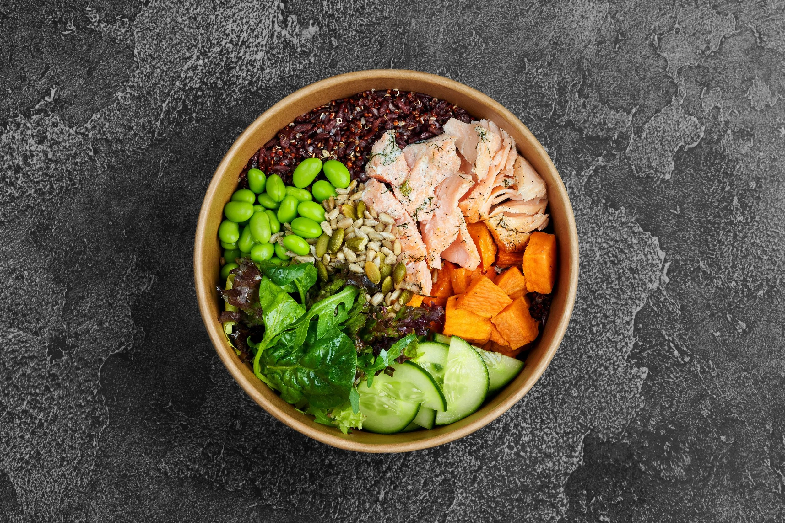 A round wooden bowl filled with mixed salad ingredients, including green edamame, sunflower seeds, shredded cooked salmon with herbs, roasted sweet potatoes, cucumber slices, and mixed greens, placed on a dark textured surface.
