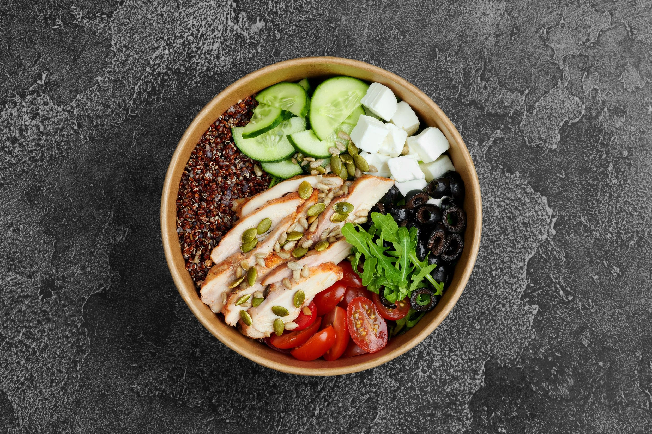 A round bowl containing a colorful salad with sliced cucumbers, cherry tomatoes, black olives, sliced grilled chicken, quartered white cheese, red quinoa, sunflower seeds, pumpkin seeds, and arugula on a dark, textured surface.