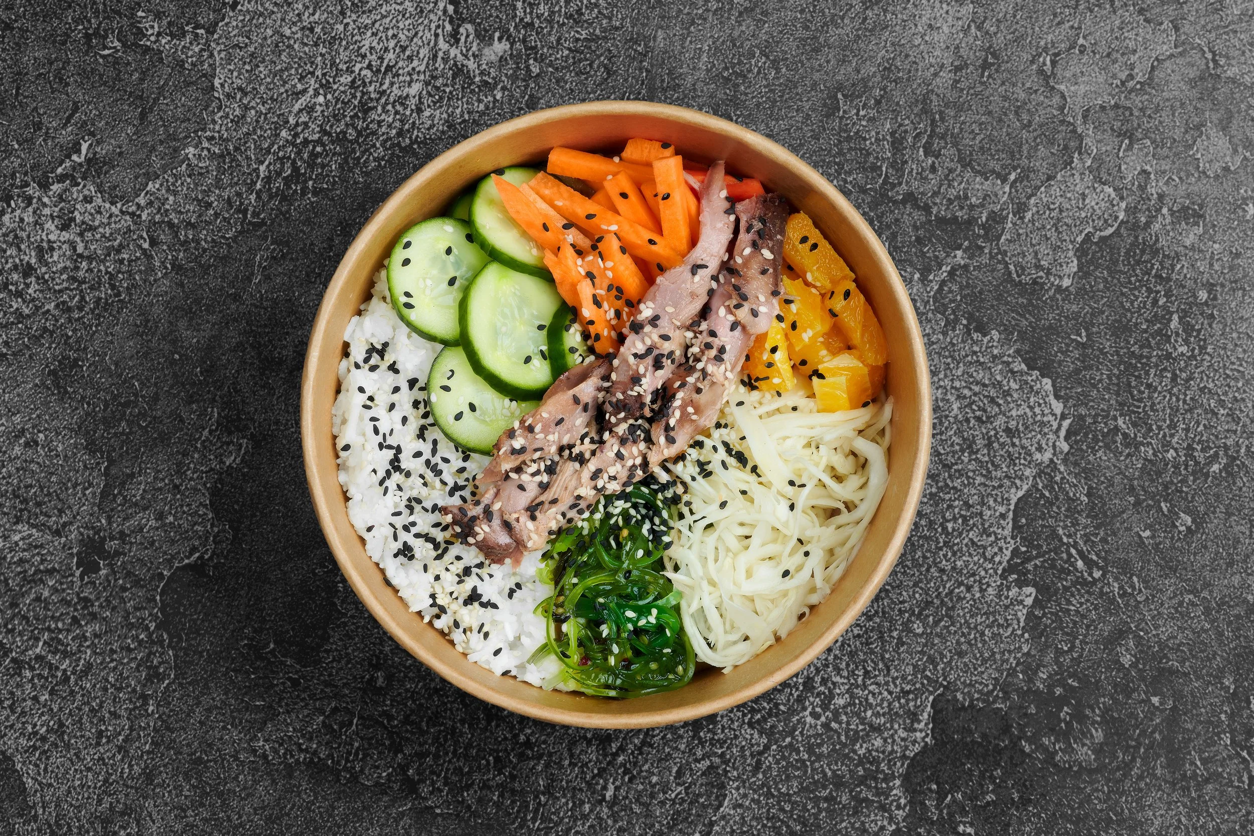 A bowl of sushi with various fresh vegetables, fish, and rice, topped with sesame seeds, on a dark textured surface.