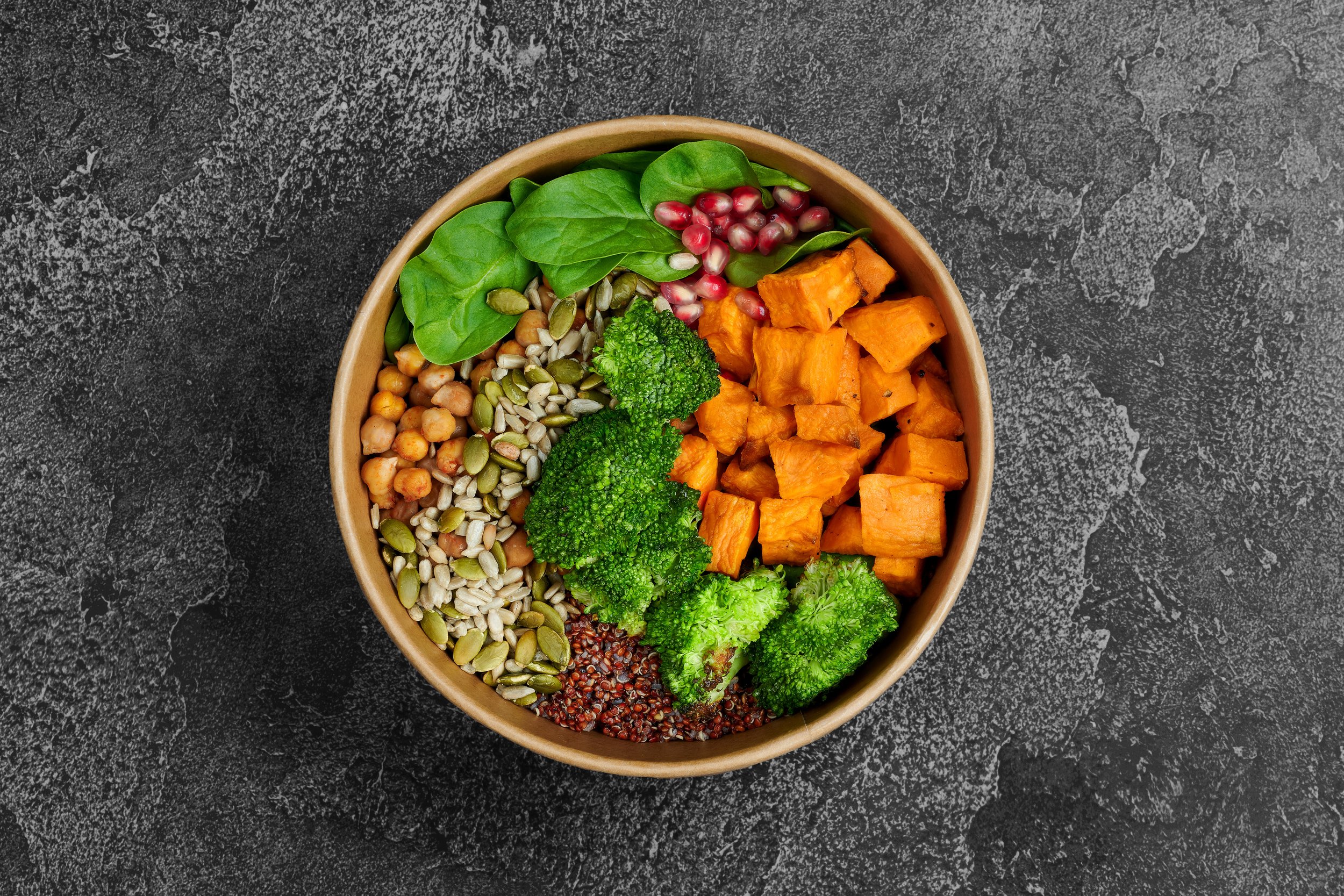 A round wooden bowl filled with a variety of fresh vegetables including spinach, broccoli, sweet potatoes, pumpkin seeds, chickpeas, and red quinoa, placed on a dark, textured surface.