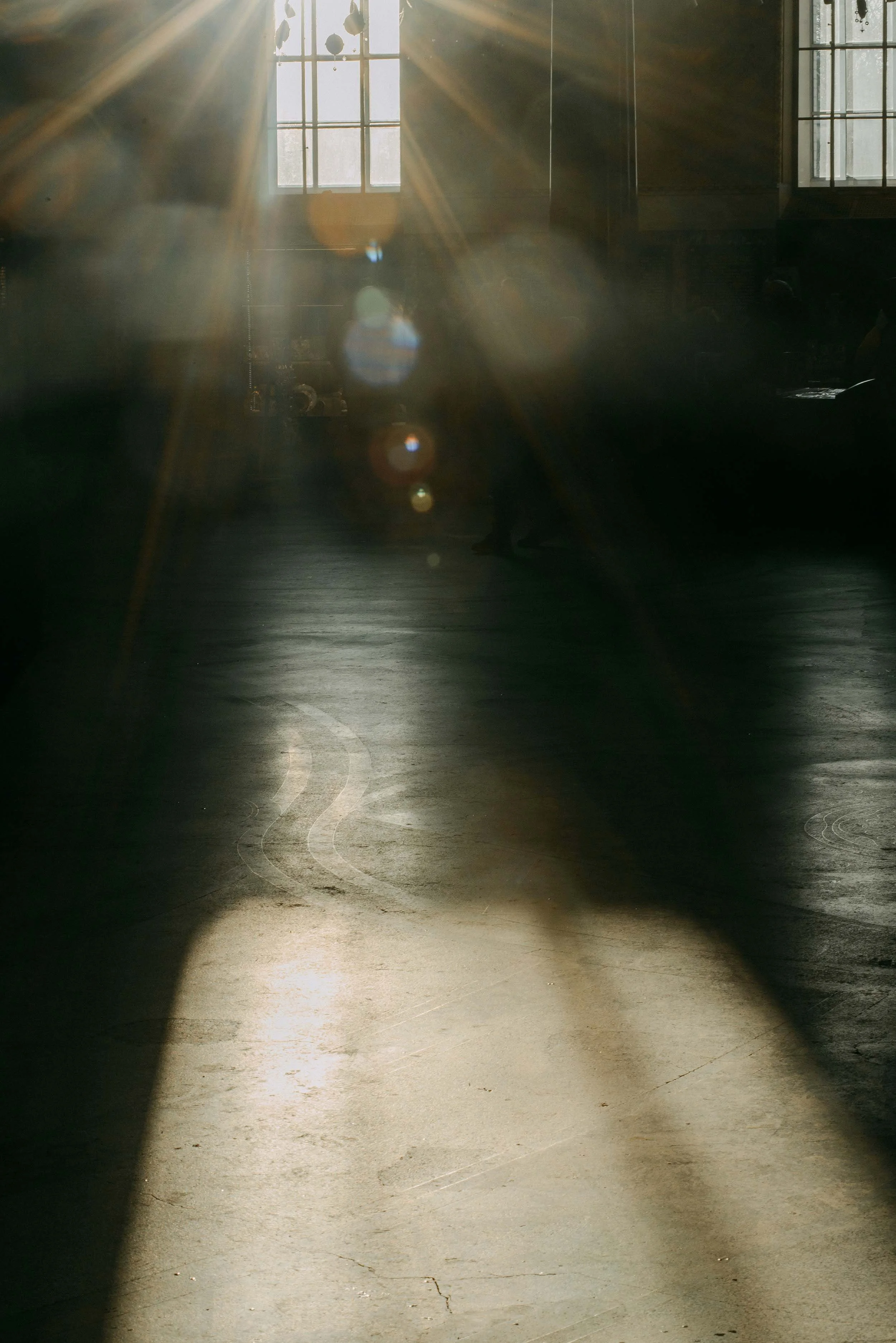 Bright sunlight streaming through large windows in an industrial-style interior, creating lens flare and reflections on a concrete floor.