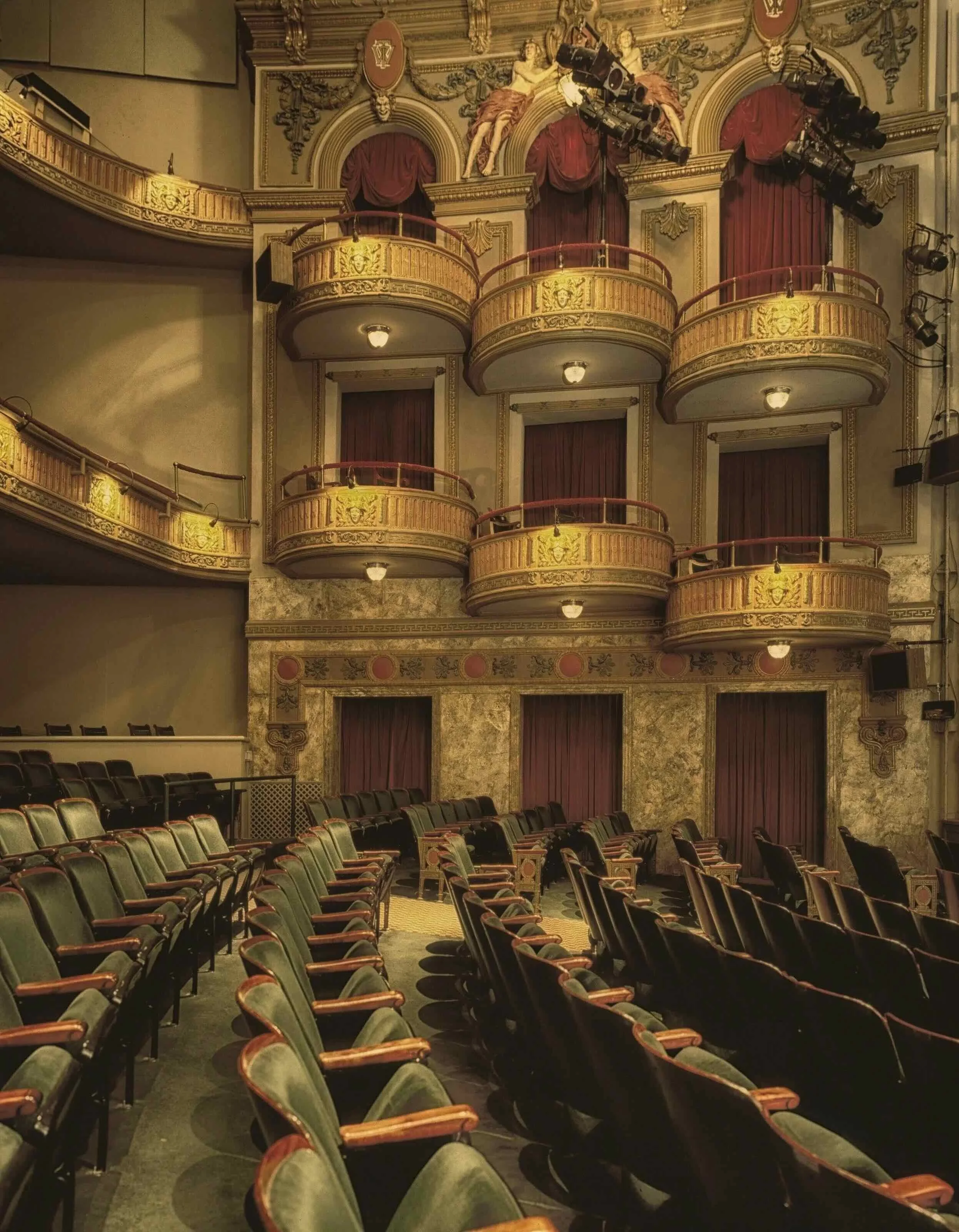 Empty theater with rows of green and black seats, ornate gold balconies and red velvet curtains in the background.