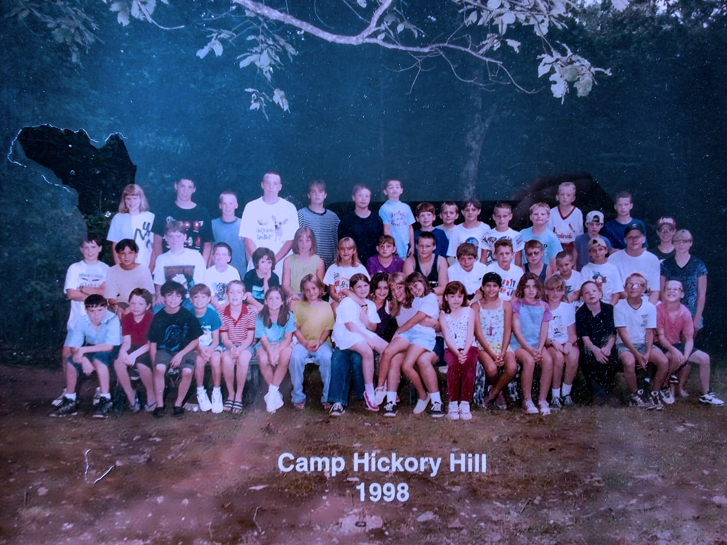 A group of children and adults posing for a photo at Camp Hickory Hill in 1998. They are outdoors, surrounded by trees, with a dirt ground in front. The group is arranged in several rows, with children seated in the front and adults standing in the back.