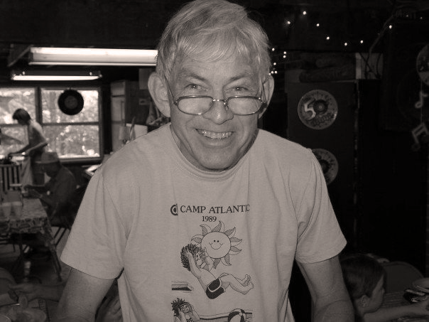 A smiling man wearing glasses and a Camp Atlantic t-shirt, standing in a room with other people, musical decorations, and windows in the background.
