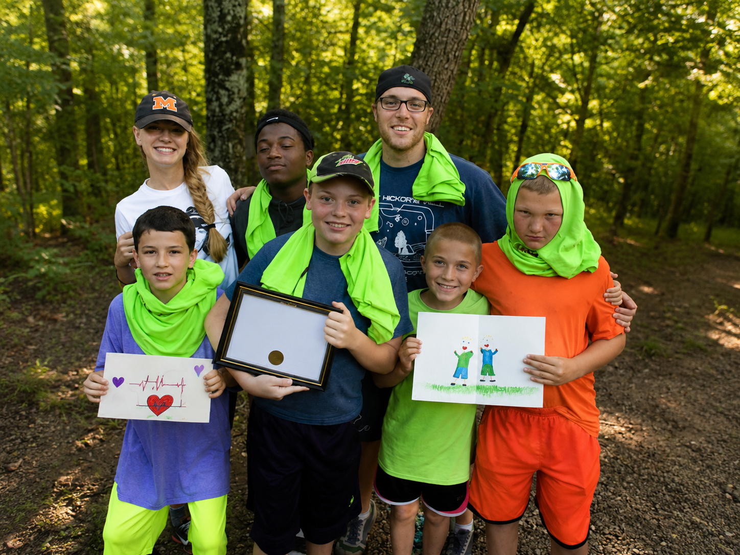 Group of children and two adults smiling in a forest, some holding drawings and a certificate.
