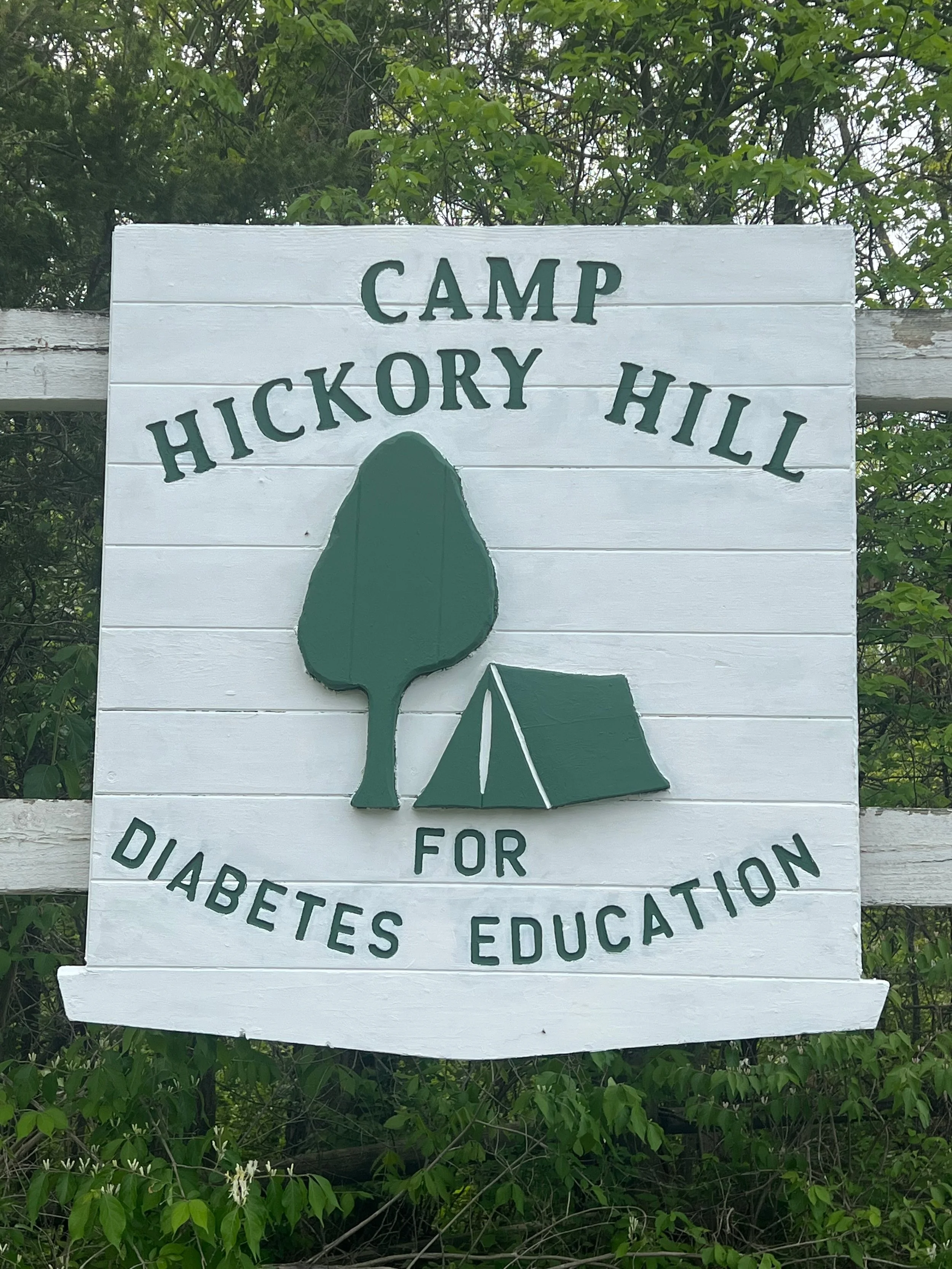 A white painted wooden sign with green text and illustrations, reading 'Camp Hickory Hill for Diabetes Education'. The sign features a green tree and a small tent illustration, and is mounted on a white fence with green foliage in the background.