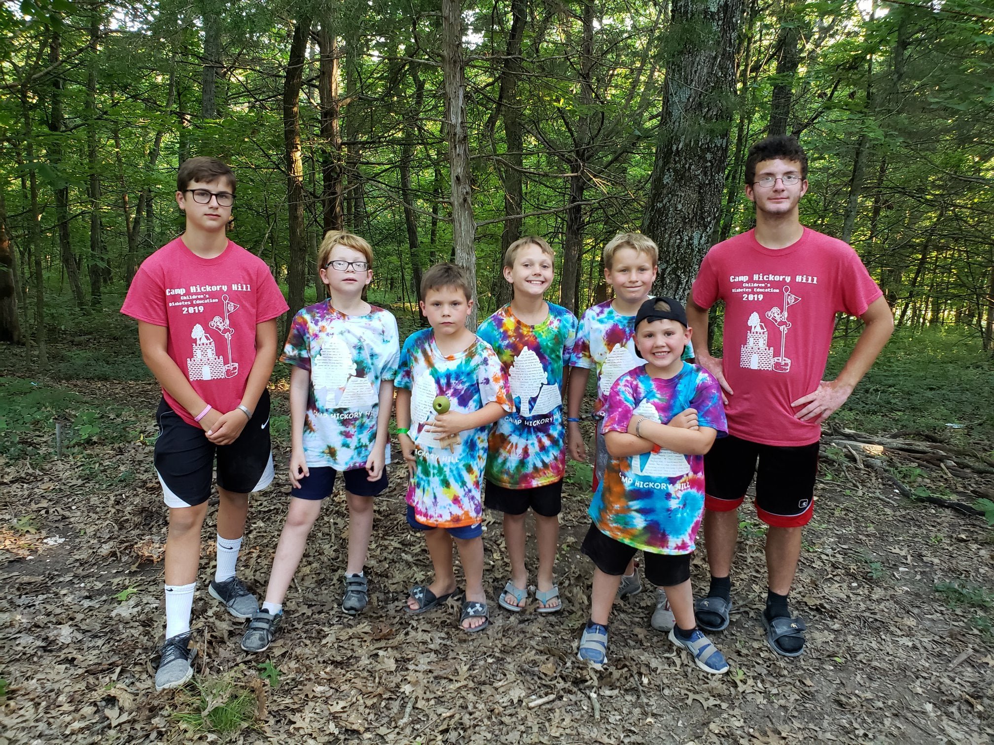 Group of young boys and a young man standing outdoors in a wooded area, some wearing tie-dye shirts, some holding paper crafts, with trees and forest background.