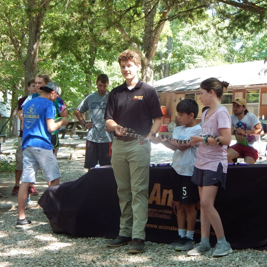 Group of children and a young man holding a large snake outdoors under trees at a daytime event.
