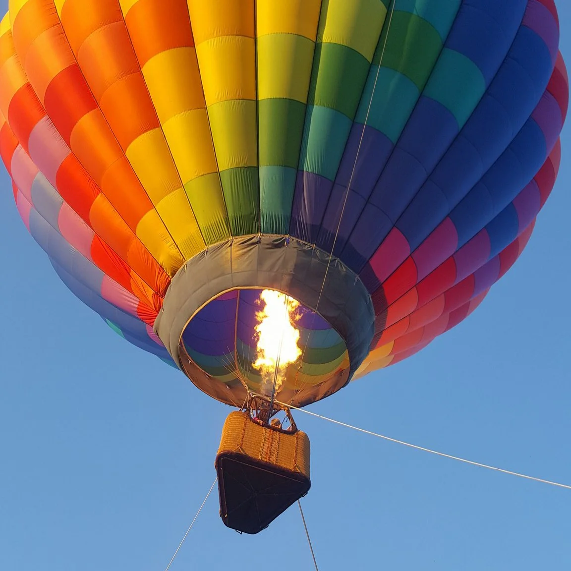 Colorful hot air balloon with rainbow pattern, inflating flame heating the air inside, flying in clear blue sky.