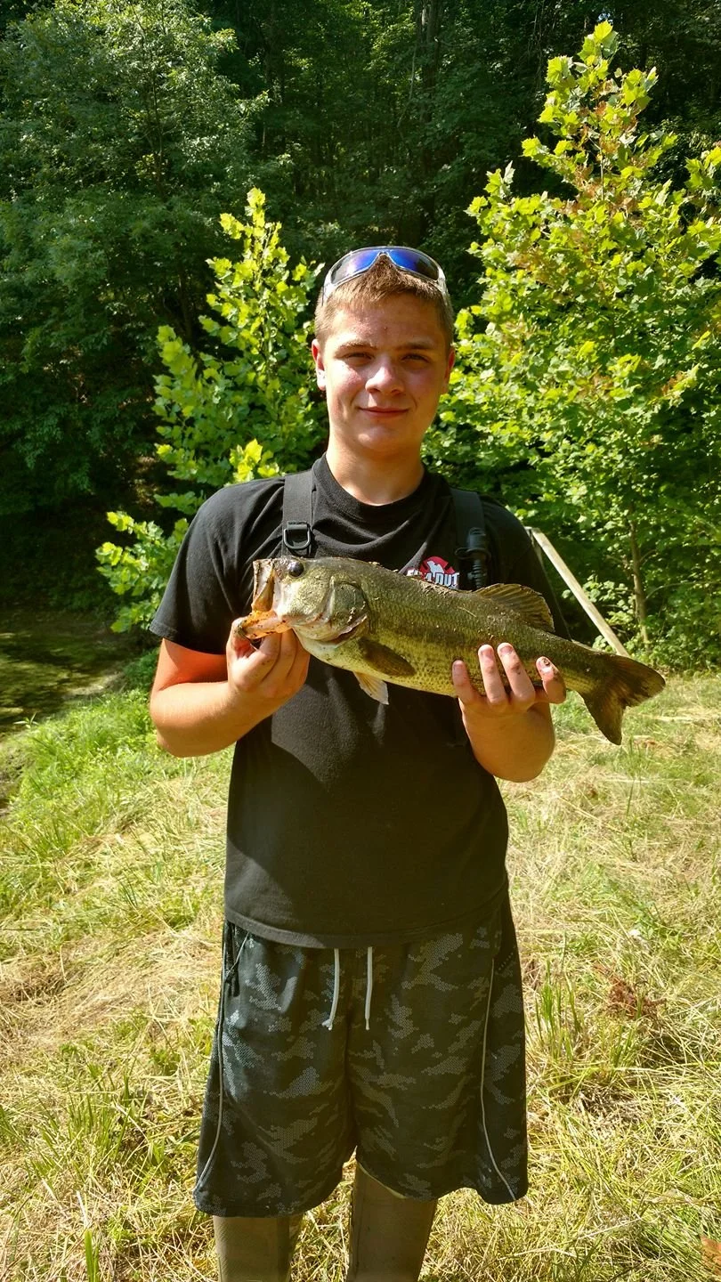 A boy holding a large fish outdoors near a forested area with sunlight shining down.