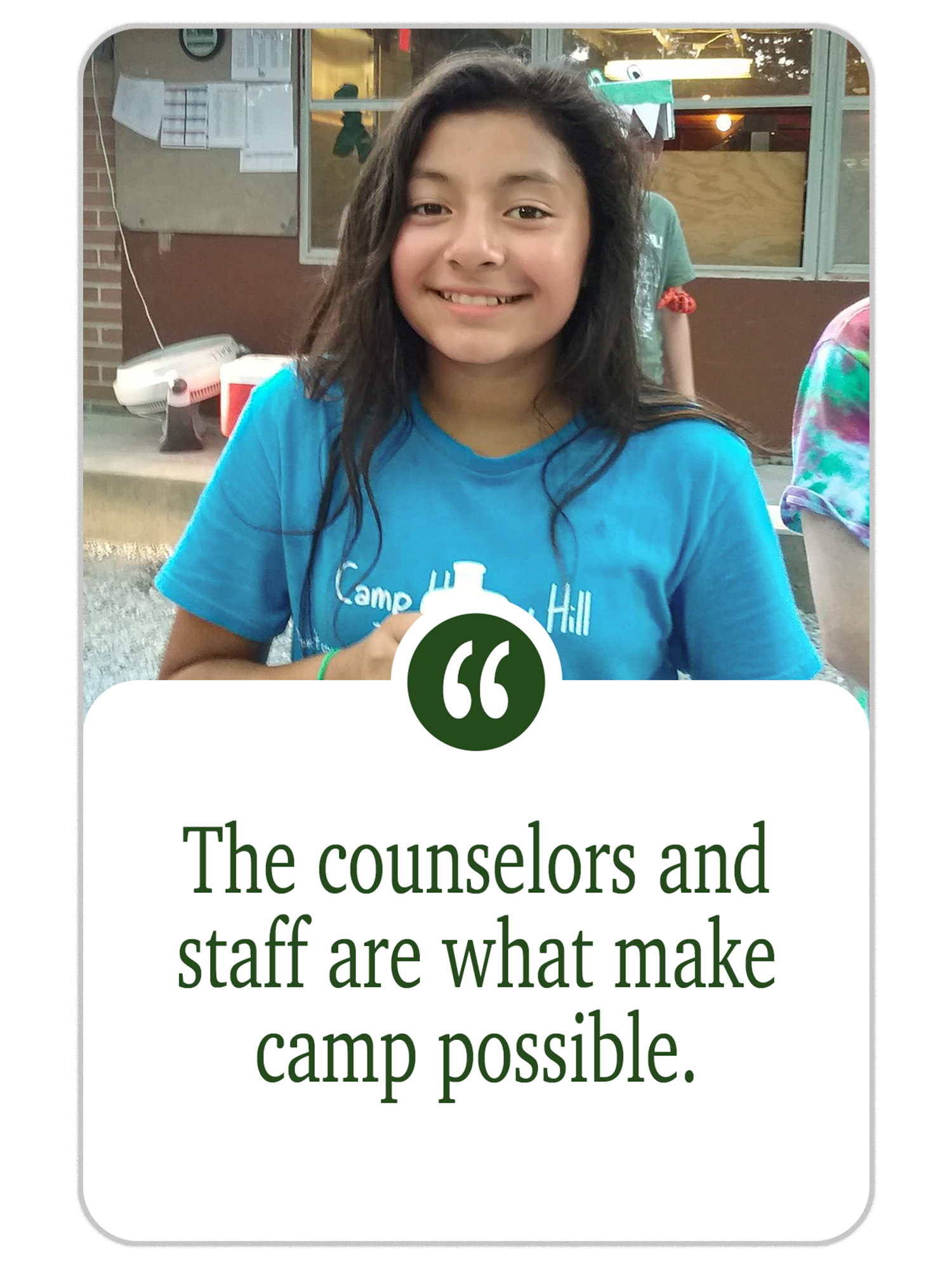 A young girl with long dark hair, smiling and wearing a blue camp shirt, sitting outdoors near a brick building with bulletin boards and a window. There is a person in a green shirt behind her.