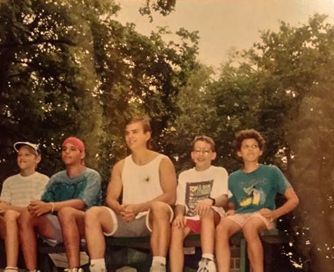 Group of five young boys sitting on a park bench outdoors among trees.