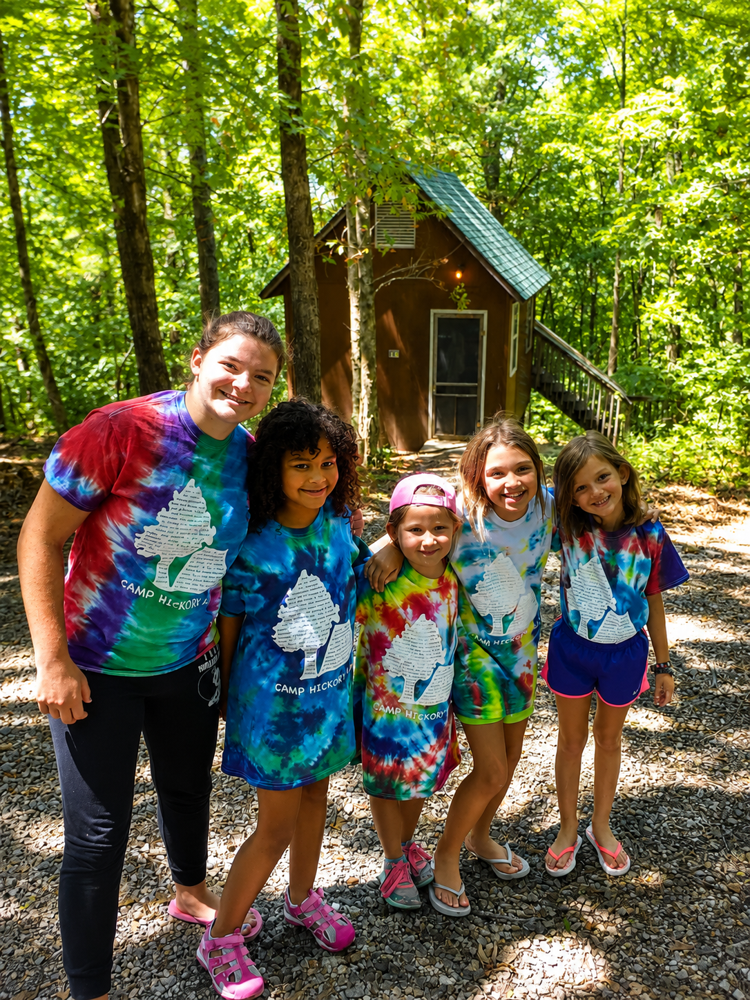 Group of five young girls smiling and standing in a wooded area in front of a small wooden cabin, all wearing colorful tie-dye camp shirts with a tree logo and the words 'Camp Hickory'.