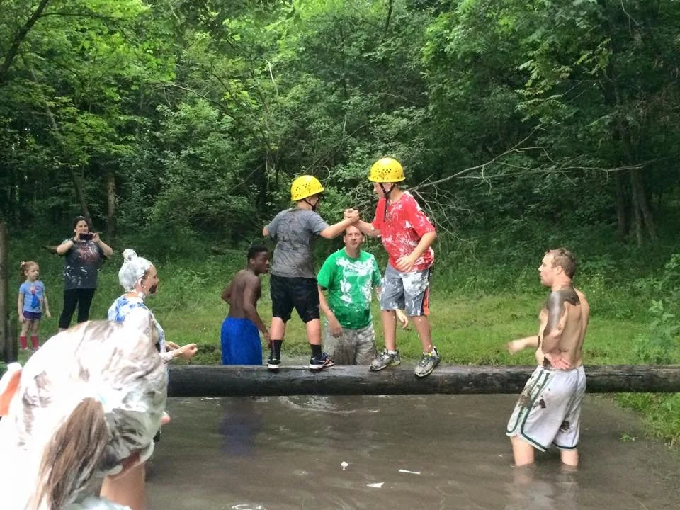 Children and adults enjoying a water activity at a creek, with some children wearing yellow protective helmets, balancing on a log across the water, surrounded by green trees.