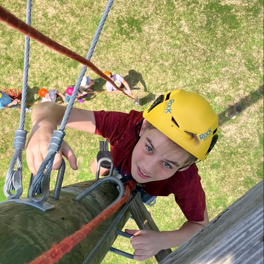 Boy climbing a wooden structure while wearing a yellow safety helmet, with a view looking up at him from below on a grassy field.
